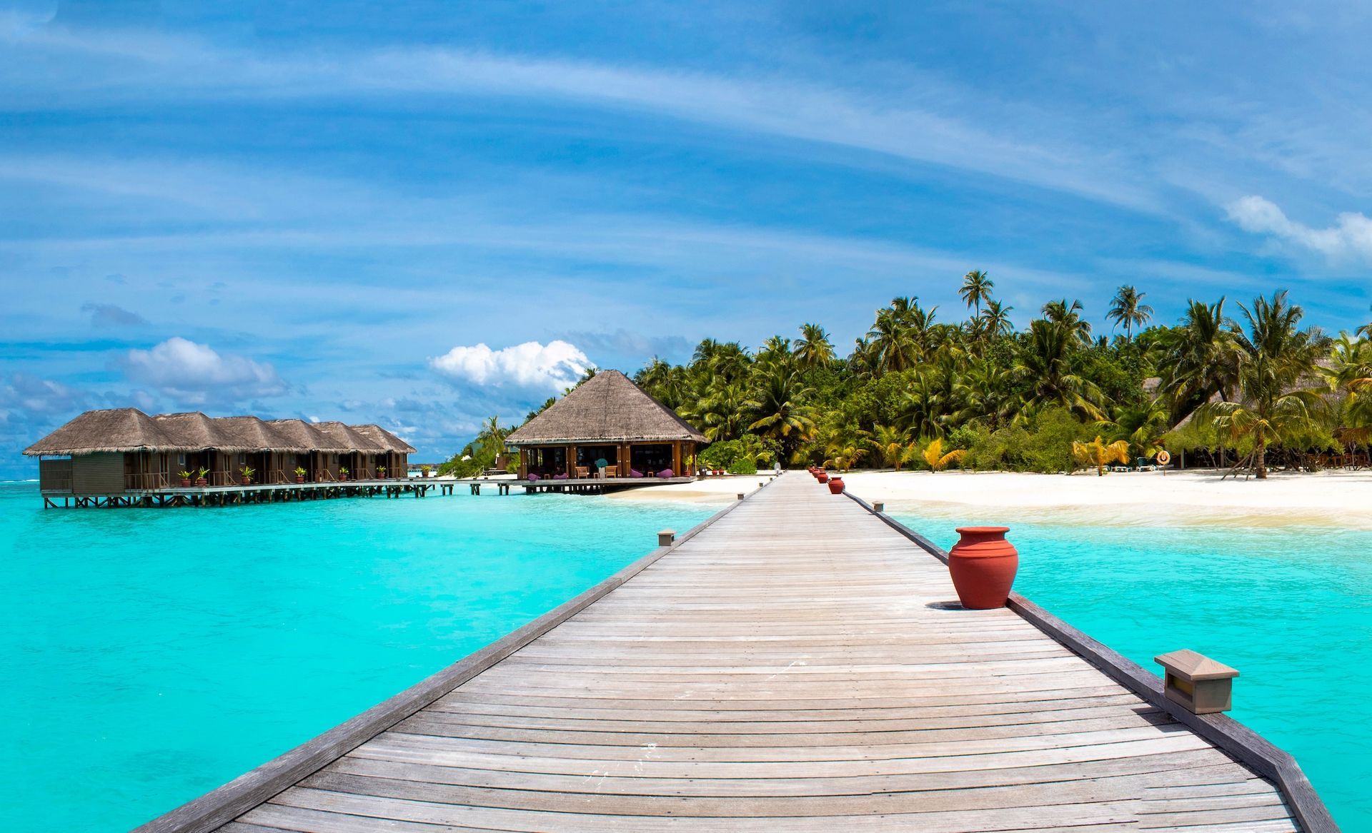 A wooden pier stretches toward thatched-roof bungalows over vibrant turquoise water on a tropical island beach.