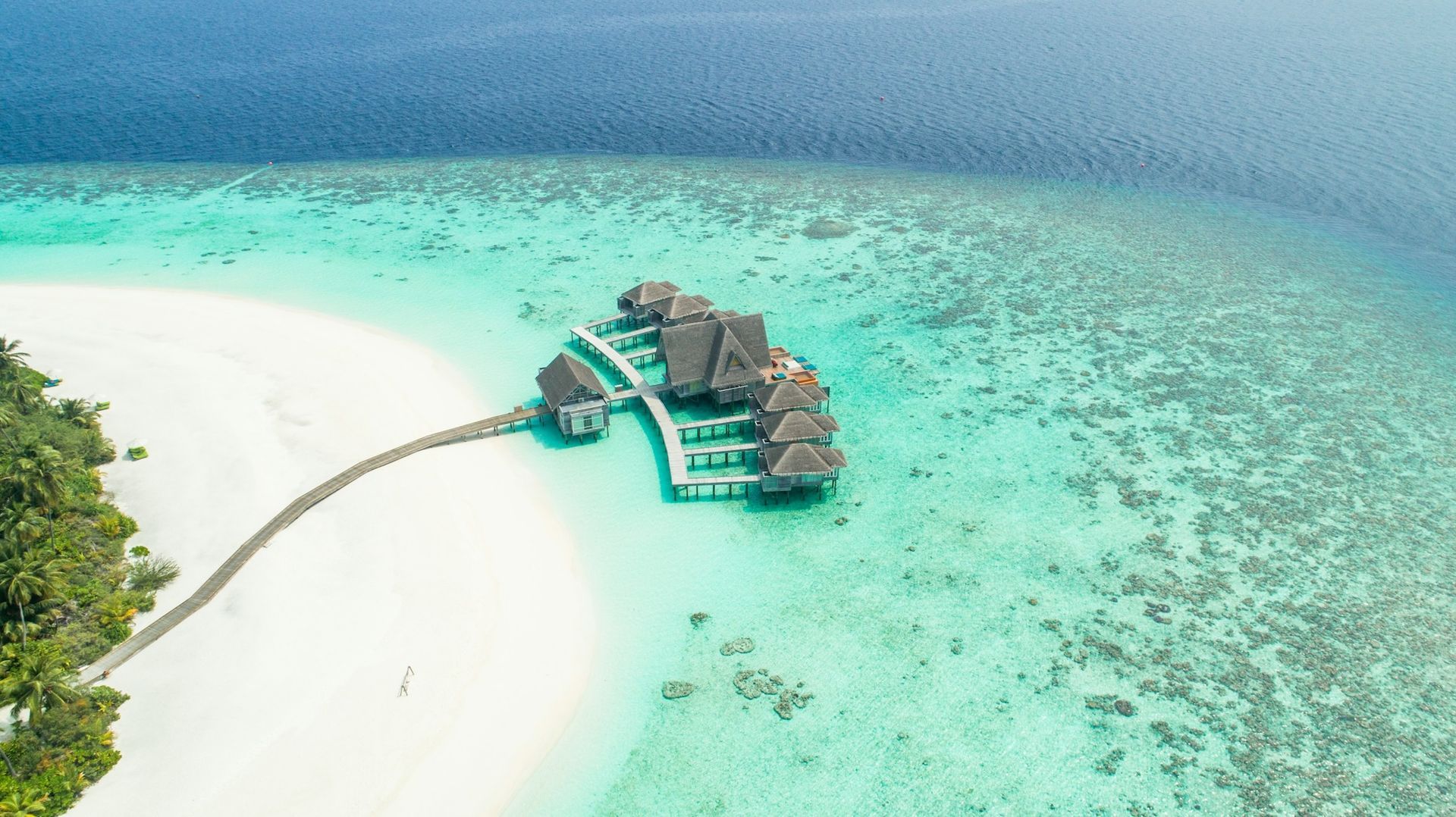 Aerial view of overwater bungalows extending from a white sand beach into clear turquoise ocean water.