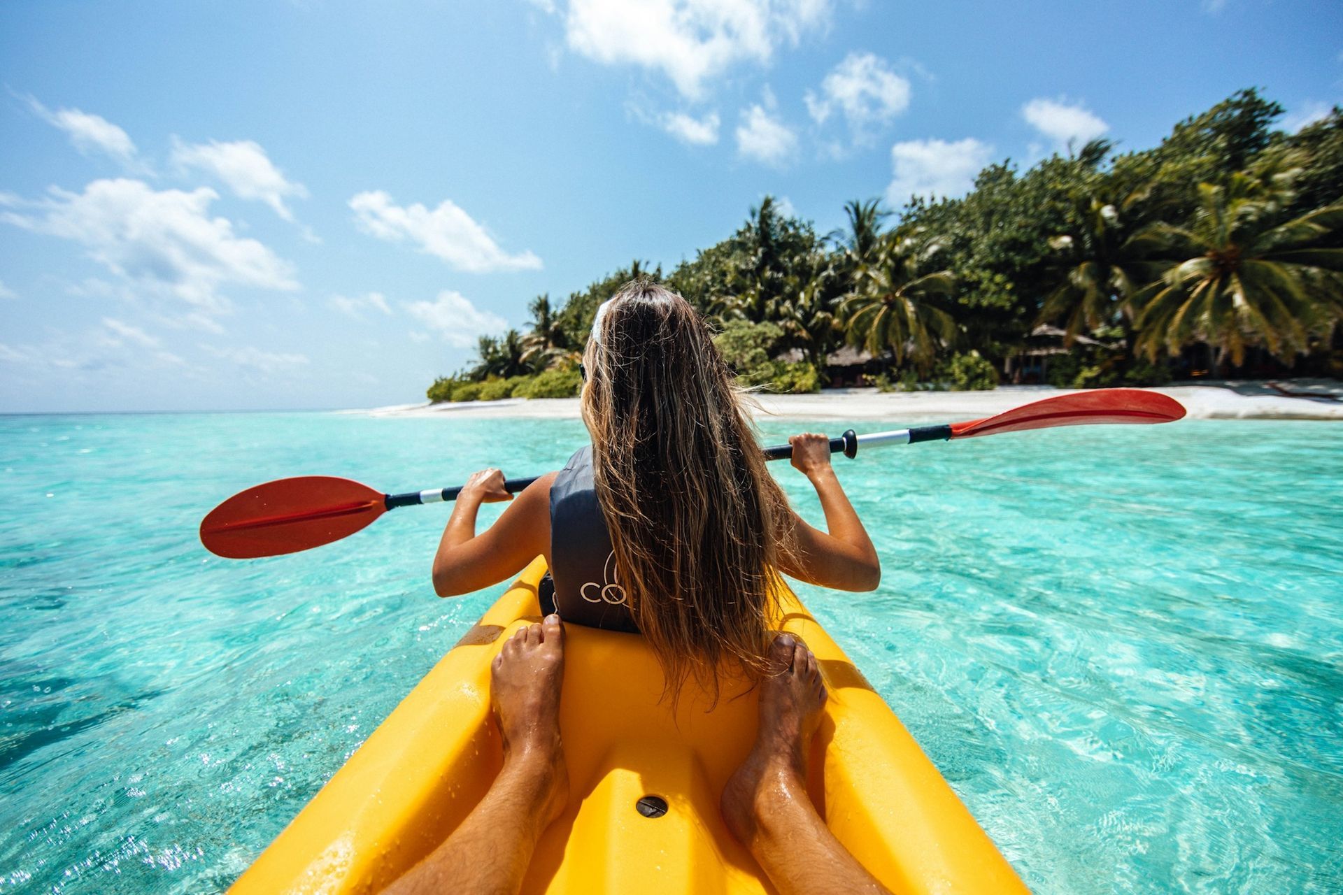 A person kayaks in clear turquoise tropical water toward a palm-fringed beach under a bright blue sky.