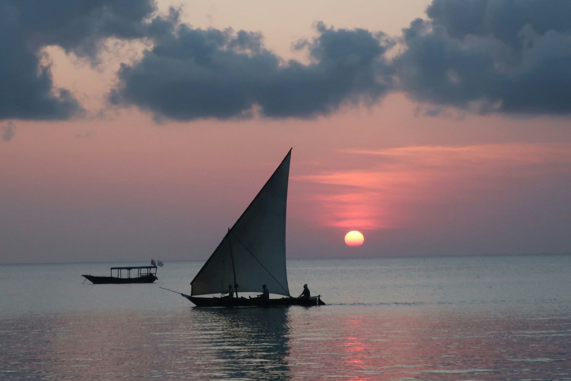 A dhow with a triangular sail floats on calm water at sunset, with a smaller motorboat in the distance.
