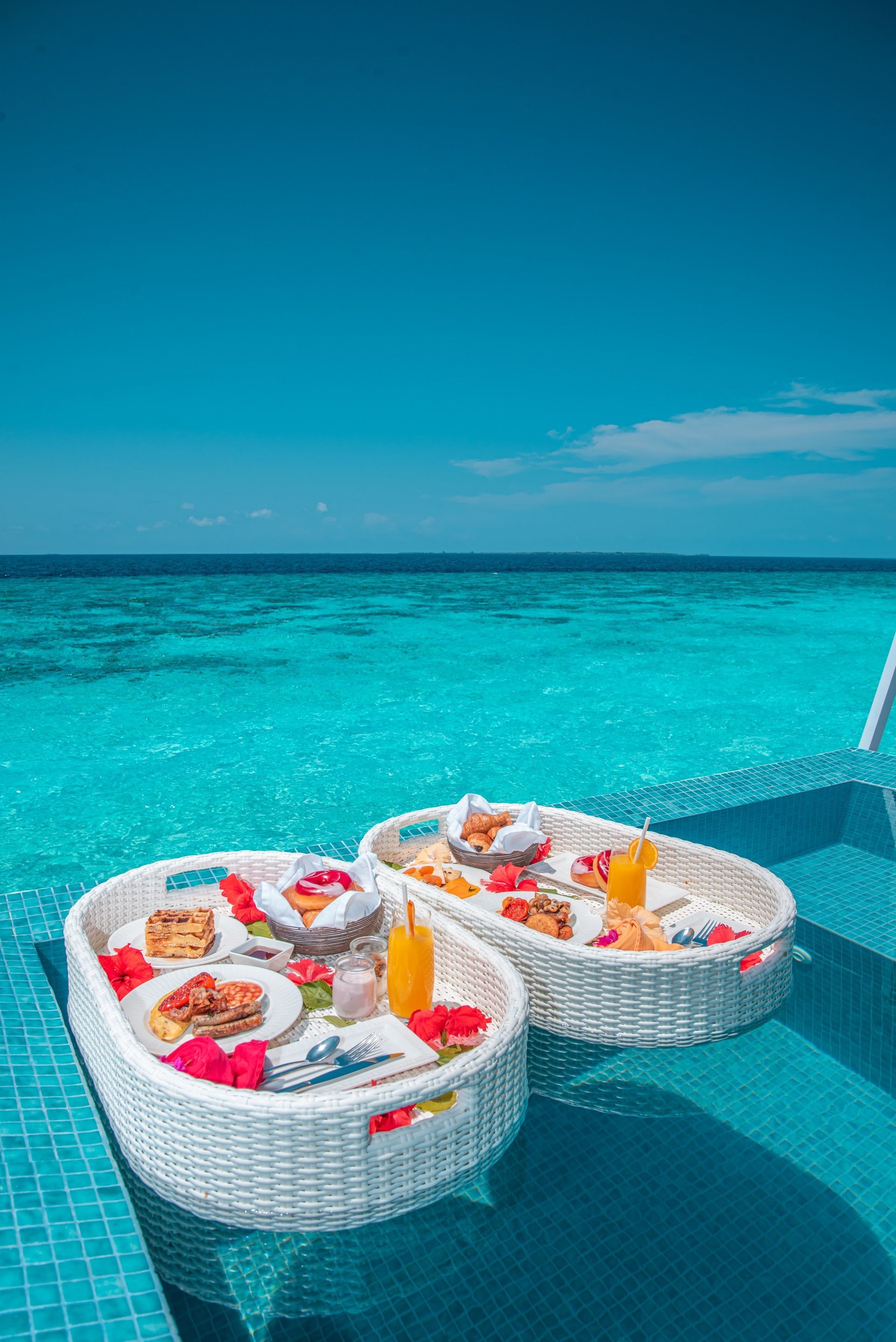 Two floating white woven baskets filled with breakfast food and juices in a swimming pool overlooking a blue ocean.