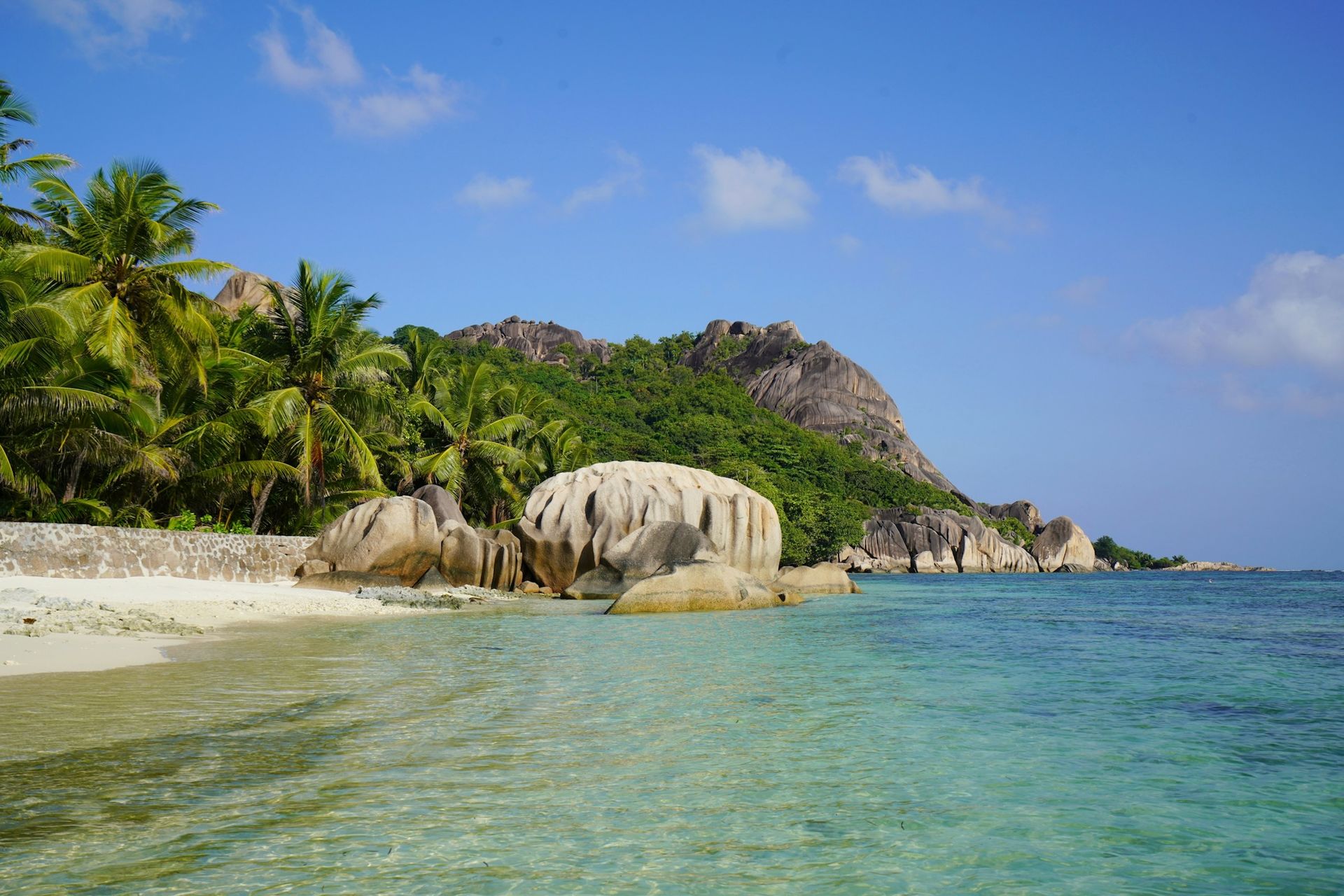 Tropical beach with large granite boulders, white sand, lush green forest, and clear turquoise water under a blue sky.