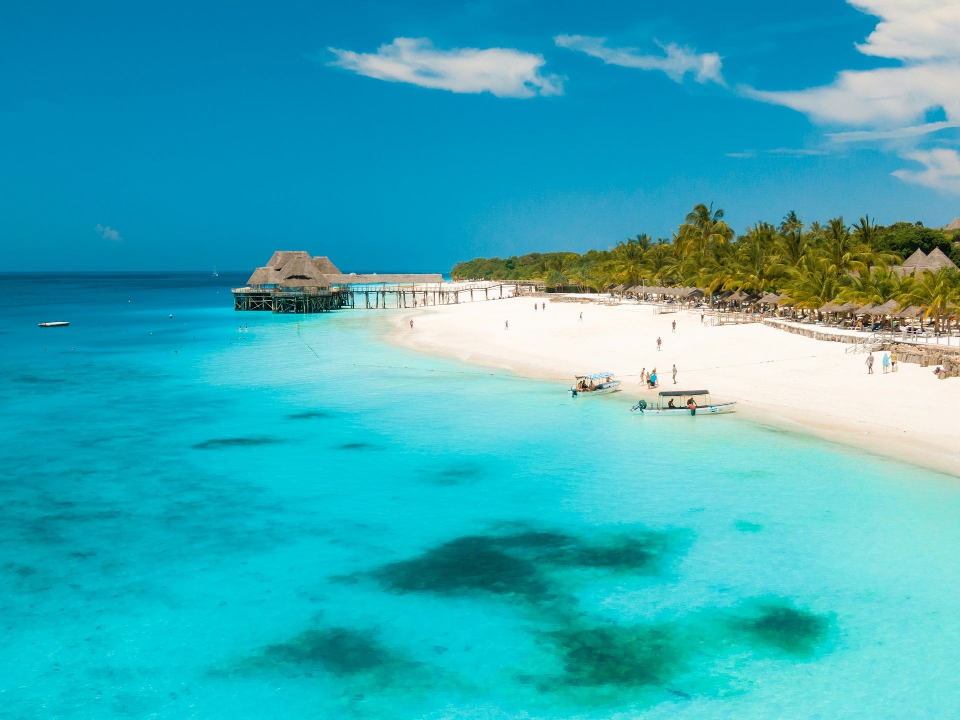 A pristine white sand beach meets turquoise water with small boats and a wooden pier leading to overwater bungalows.