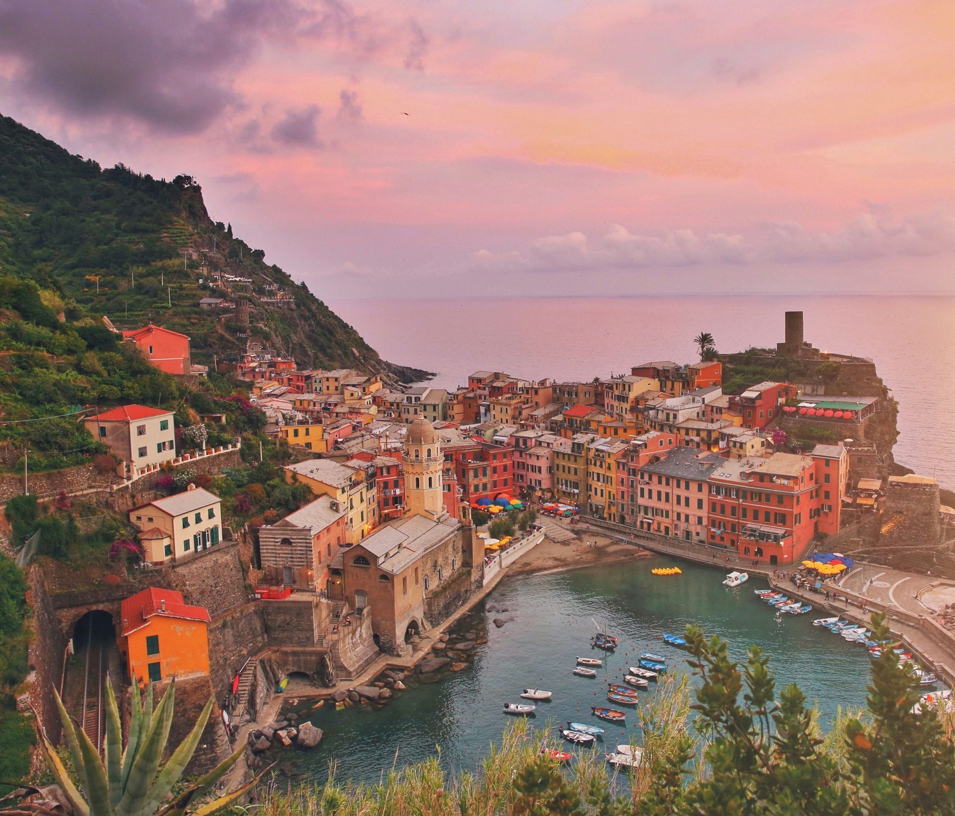 Vernazza, Italy: colorful hillside buildings overlooking a small harbor with boats at sunset.