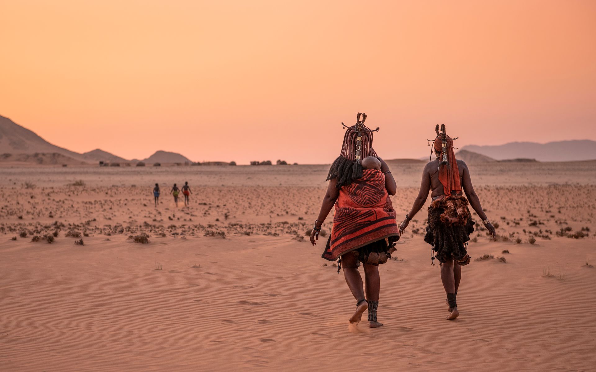 Two people in traditional, patterned attire walk across a vast, sandy desert landscape at sunset.