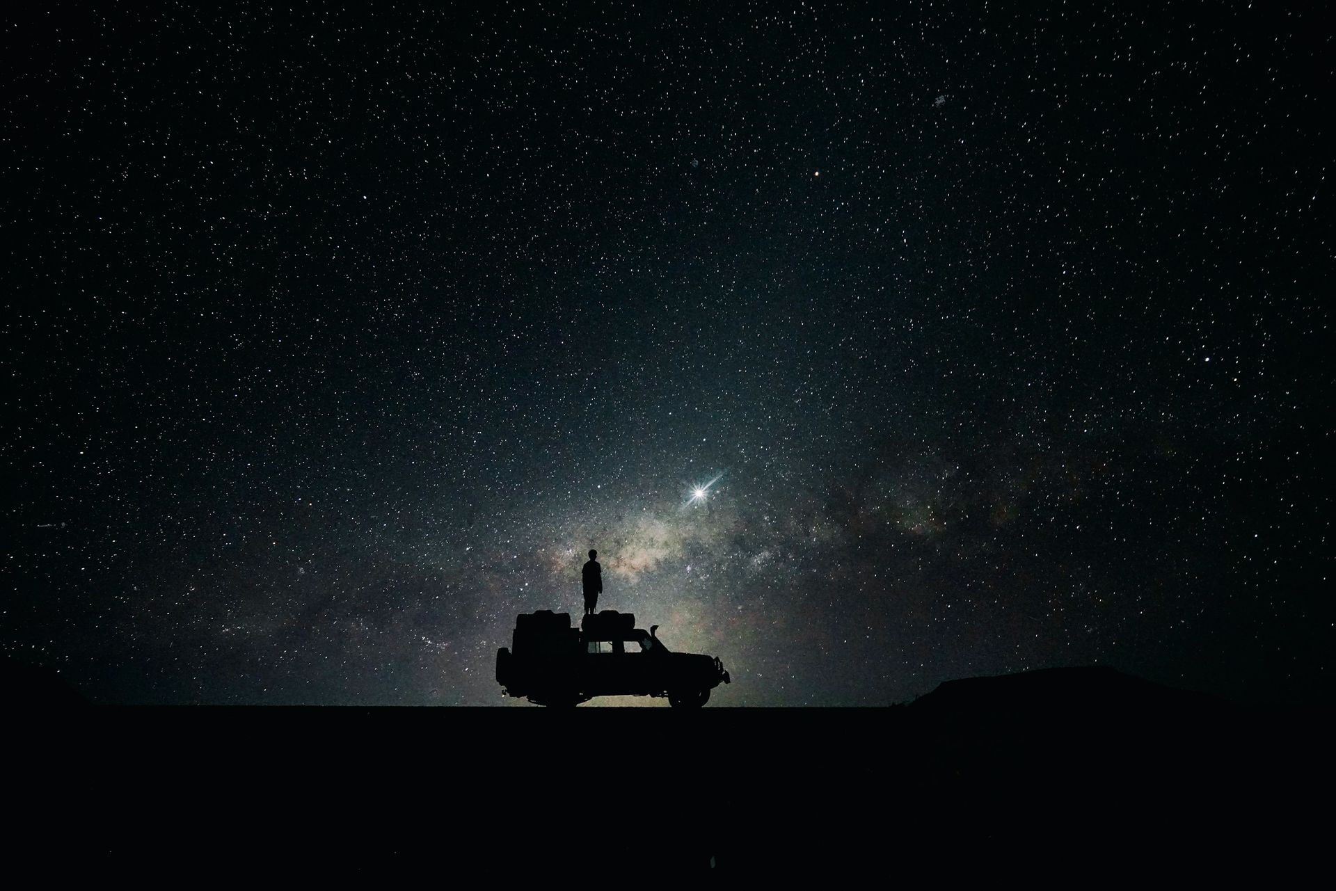 A silhouette of a person standing on top of an SUV against a starry night sky and the Milky Way.