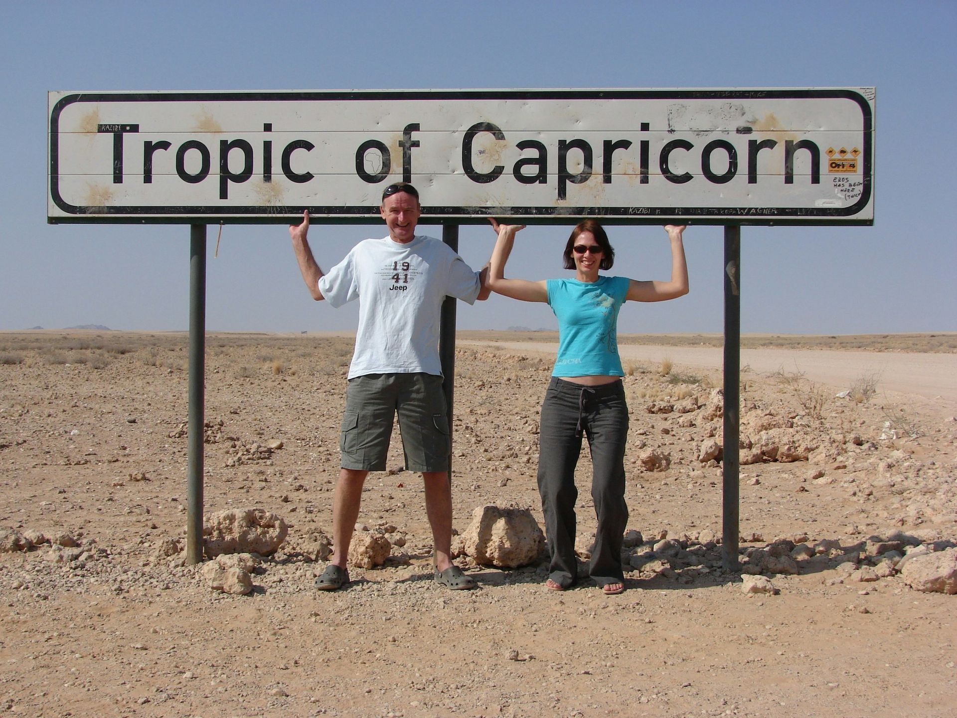 Two people stand in a desert landscape, smiling while holding up a sign that reads 