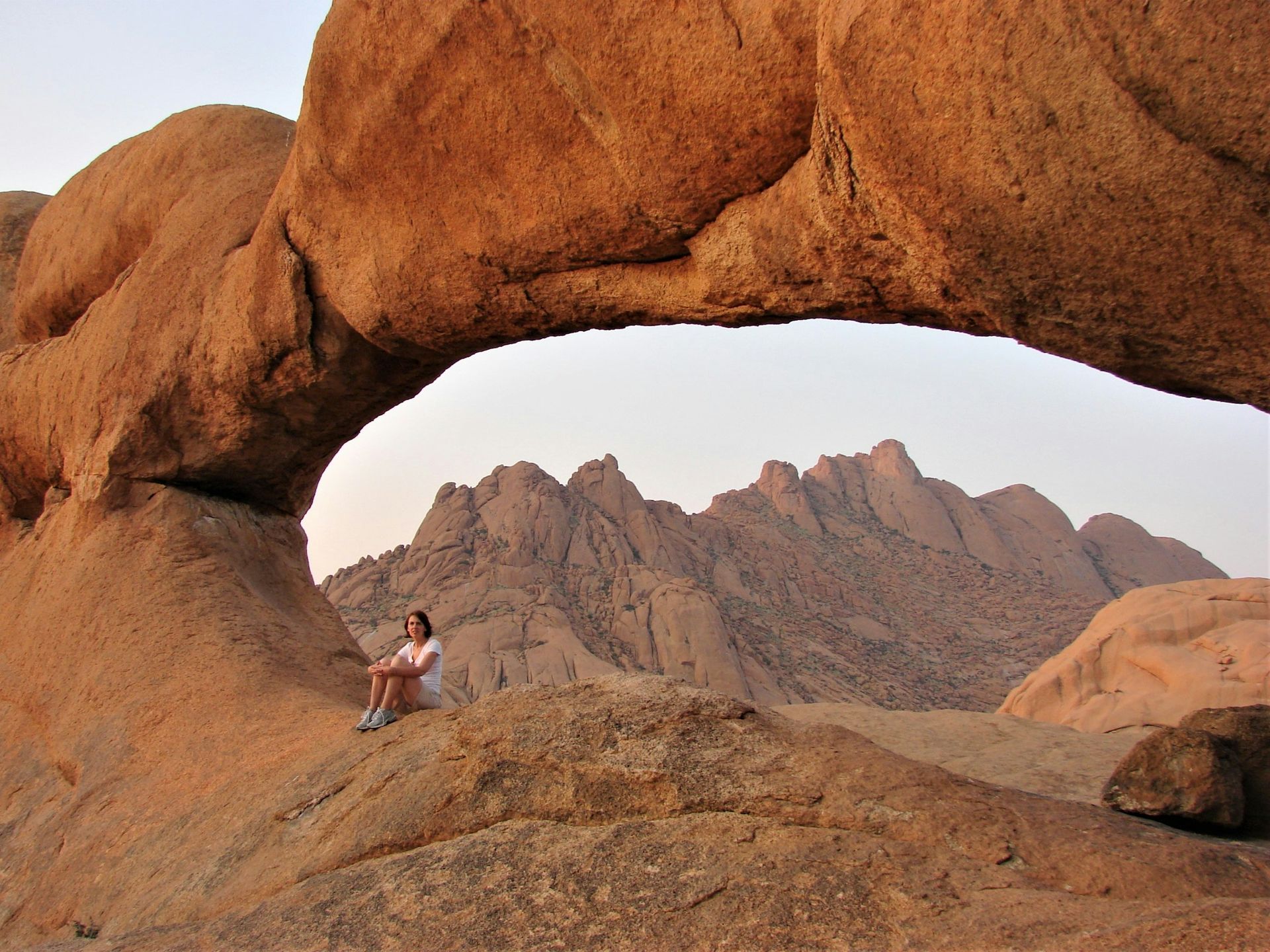 A person sits on a rock beneath a natural stone arch, looking out at a rugged mountain range in the golden afternoon light.