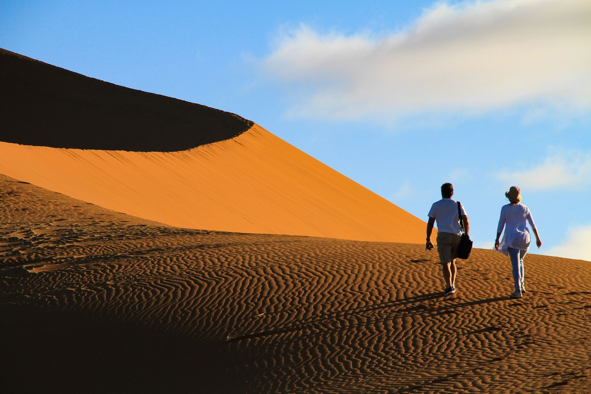 Two people walk up a rippled, orange sand dune under a blue sky with soft clouds.