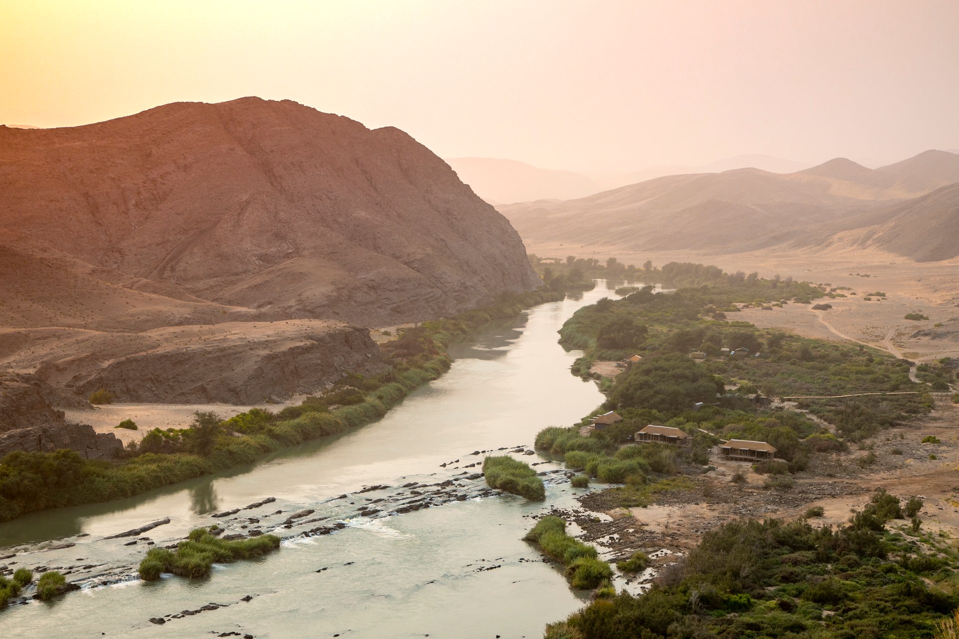 A river flows through an arid, mountainous landscape at sunset, with rocky islands and greenery lining the banks.