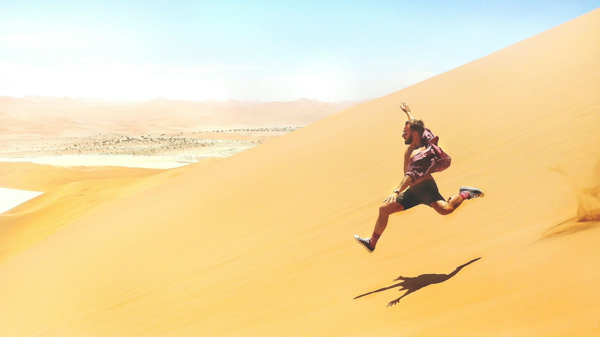 A person leaping down a steep, sunlit sand dune in a vast desert landscape.