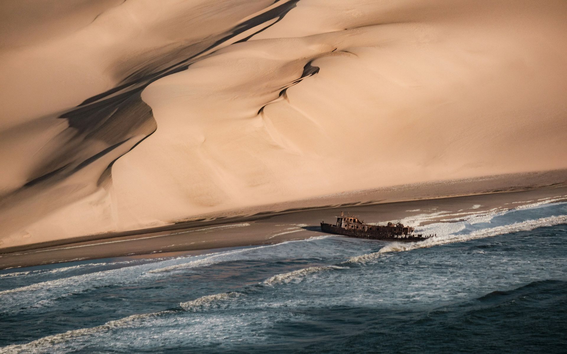 A rusted shipwreck lies on a sandy beach at the base of massive, rolling sand dunes beside the blue ocean.