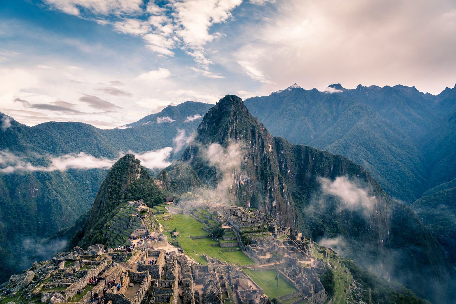 Machu Picchu ruins in the Andes mountains, surrounded by lush green peaks, mist, and a cloudy blue sky.