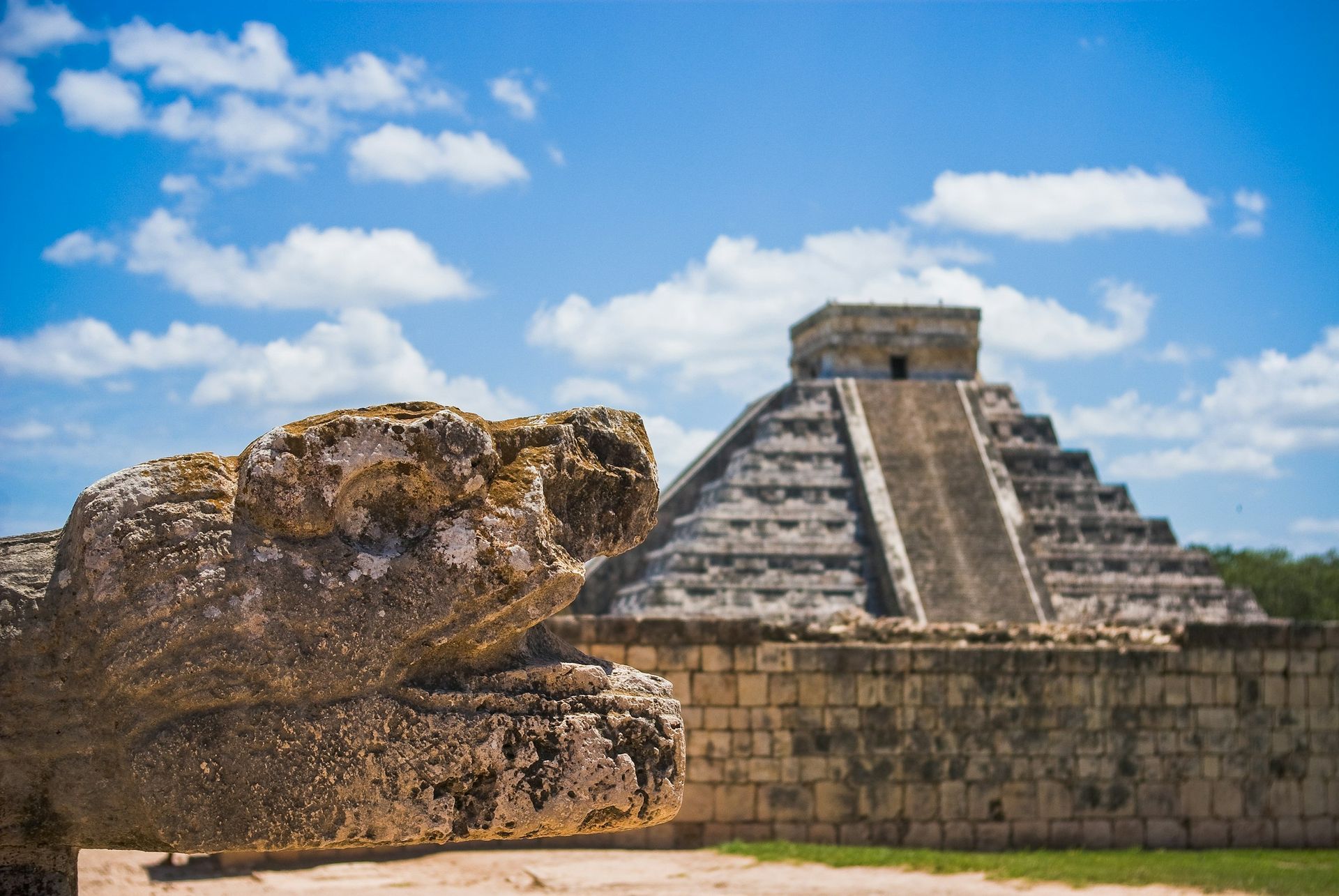 A stone serpent sculpture in the foreground with the Chichen Itza pyramid in the background under a blue, cloudy sky.