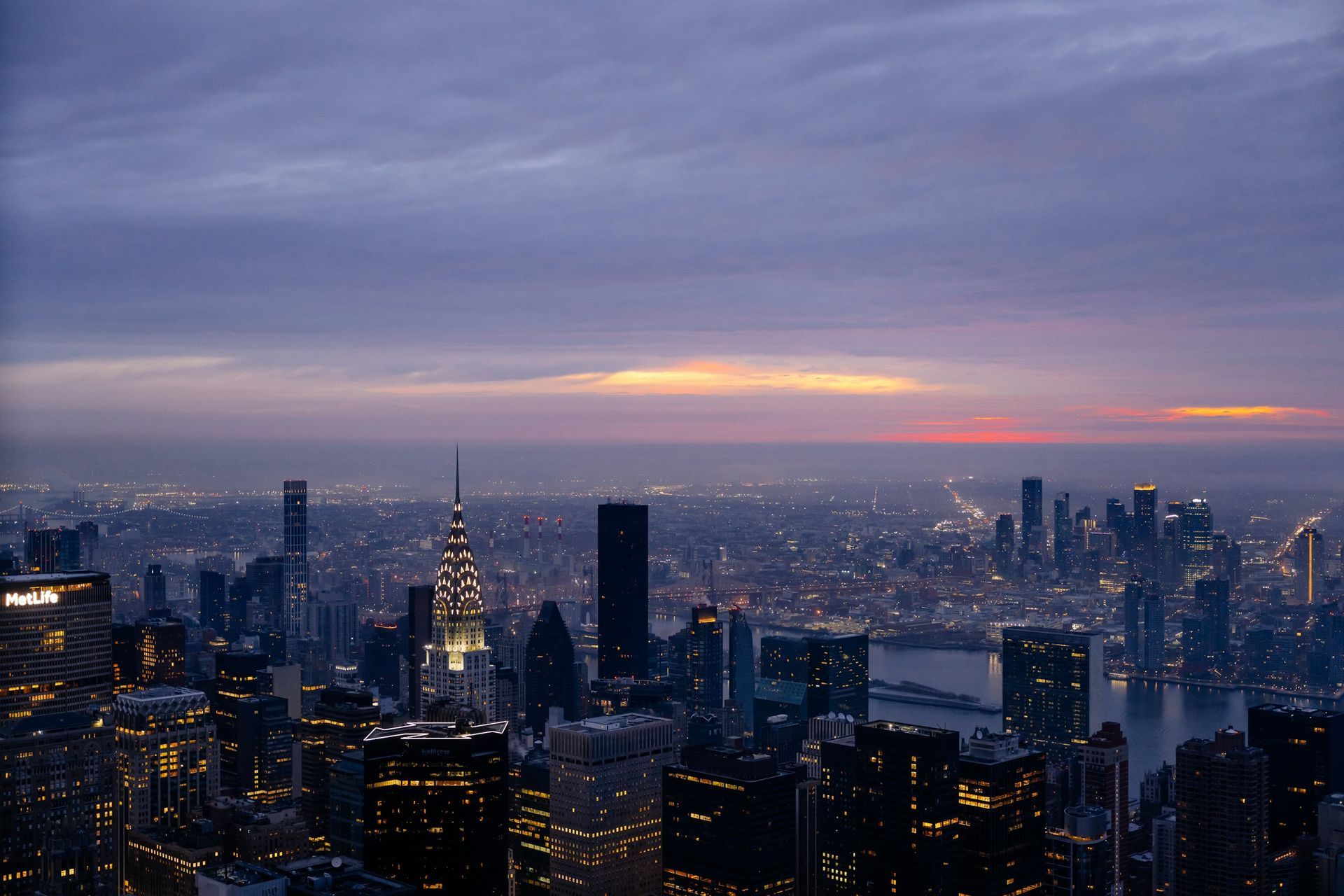 New York City skyline at dusk, featuring the illuminated Chrysler Building and distant harbor under a purple sunset.