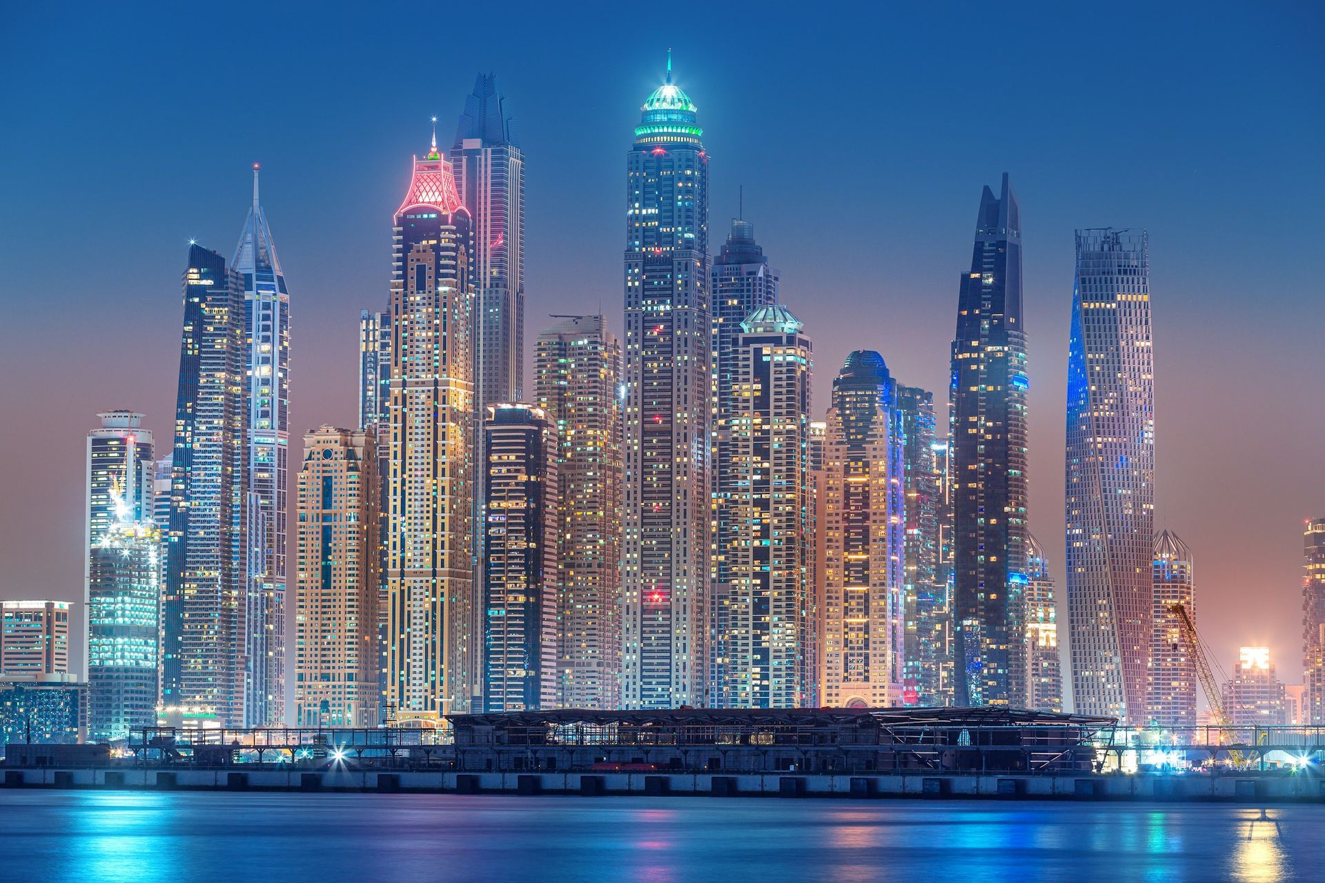 A brightly lit Dubai Marina skyline at night, featuring a cluster of modern skyscrapers reflecting over the water.