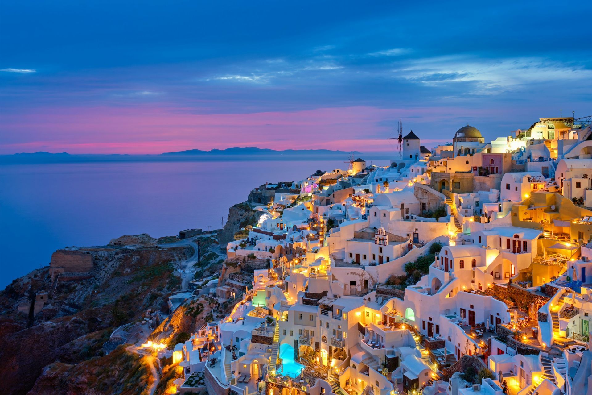 Oia, Santorini at sunset, with illuminated white buildings built into a cliffside overlooking the sea.