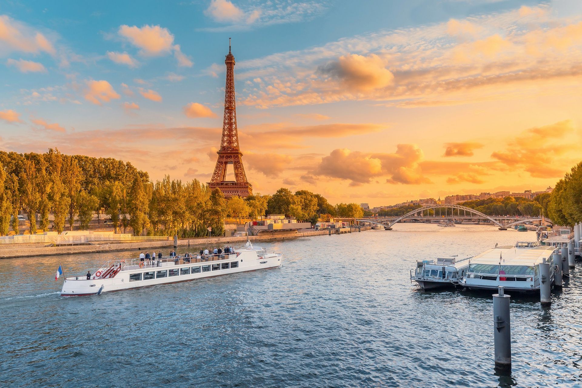 The Eiffel Tower stands above the Seine River at sunset, with a riverboat cruising past tour boats docked at the bank.