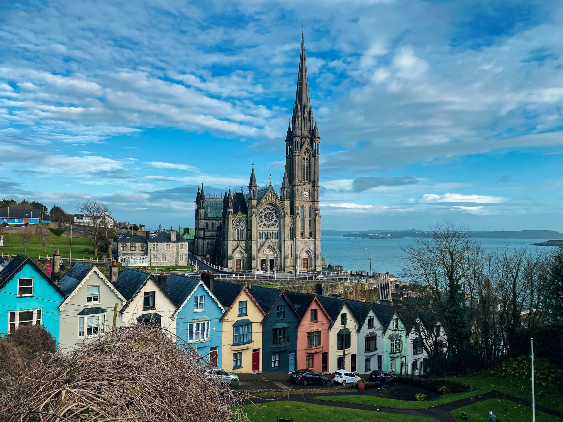A row of colorful houses below St. Colman’s Cathedral overlooking the water in Cobh, Ireland, under a cloudy blue sky.