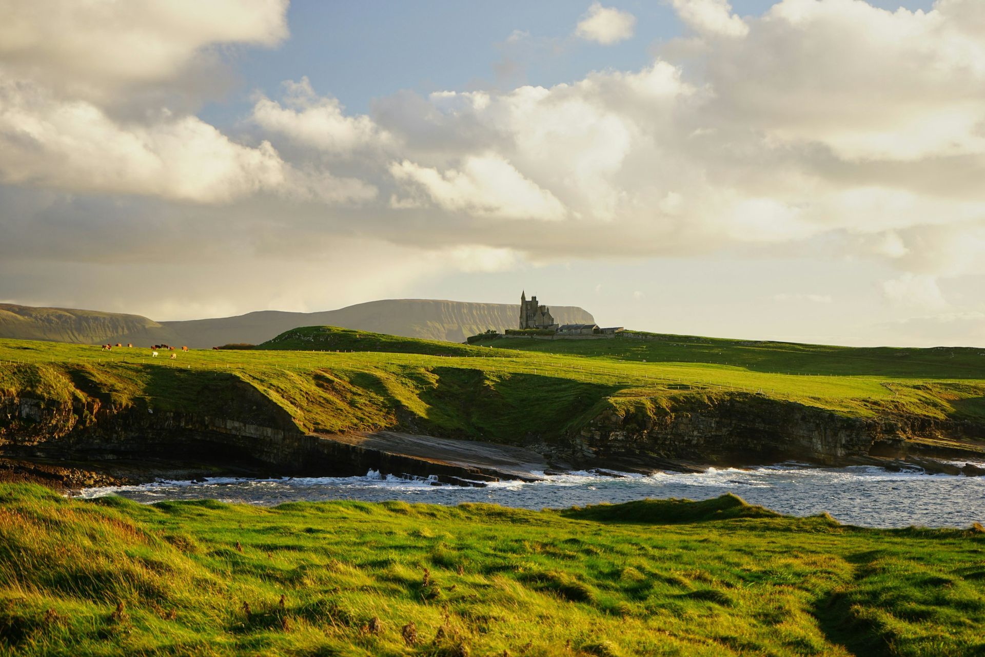 A distant, ruined stone castle stands on a green, grassy cliff above a rocky coastline under a cloudy, golden sky.