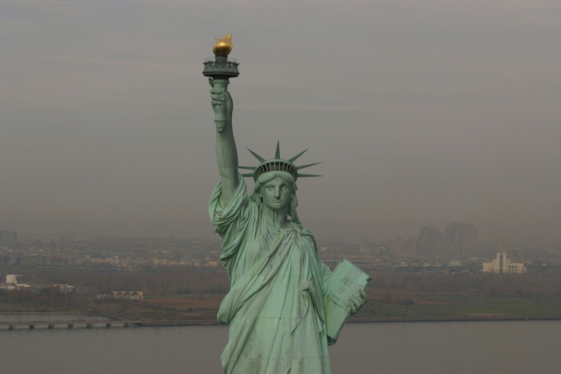 The Statue of Liberty holding a torch and tablet, set against a hazy background of water and a distant city skyline.