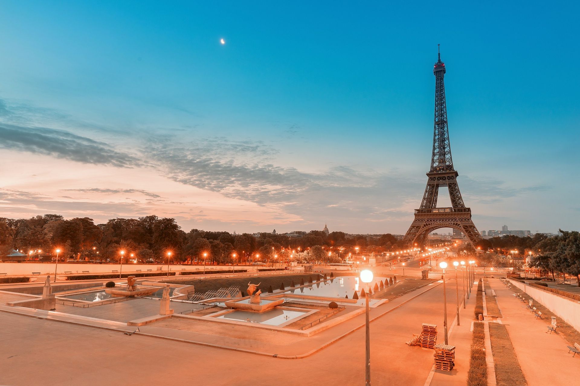 Eiffel Tower in Paris at twilight, with warm lights glowing across a plaza and a clear sky featuring a small moon.