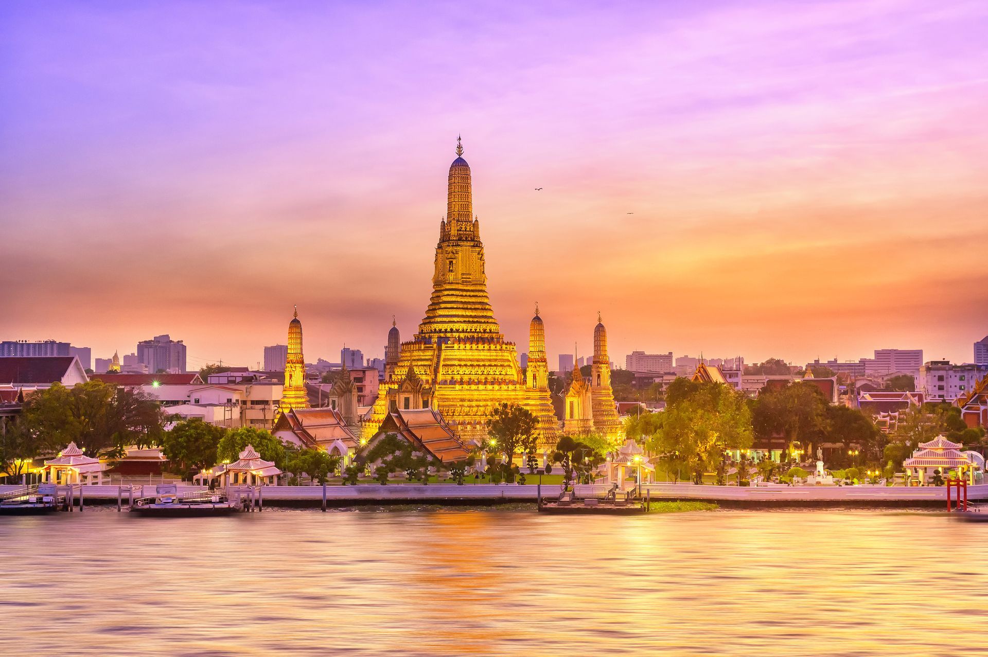 Wat Arun temple in Bangkok, Thailand, illuminated at sunset by a golden, purple sky along the Chao Phraya River.