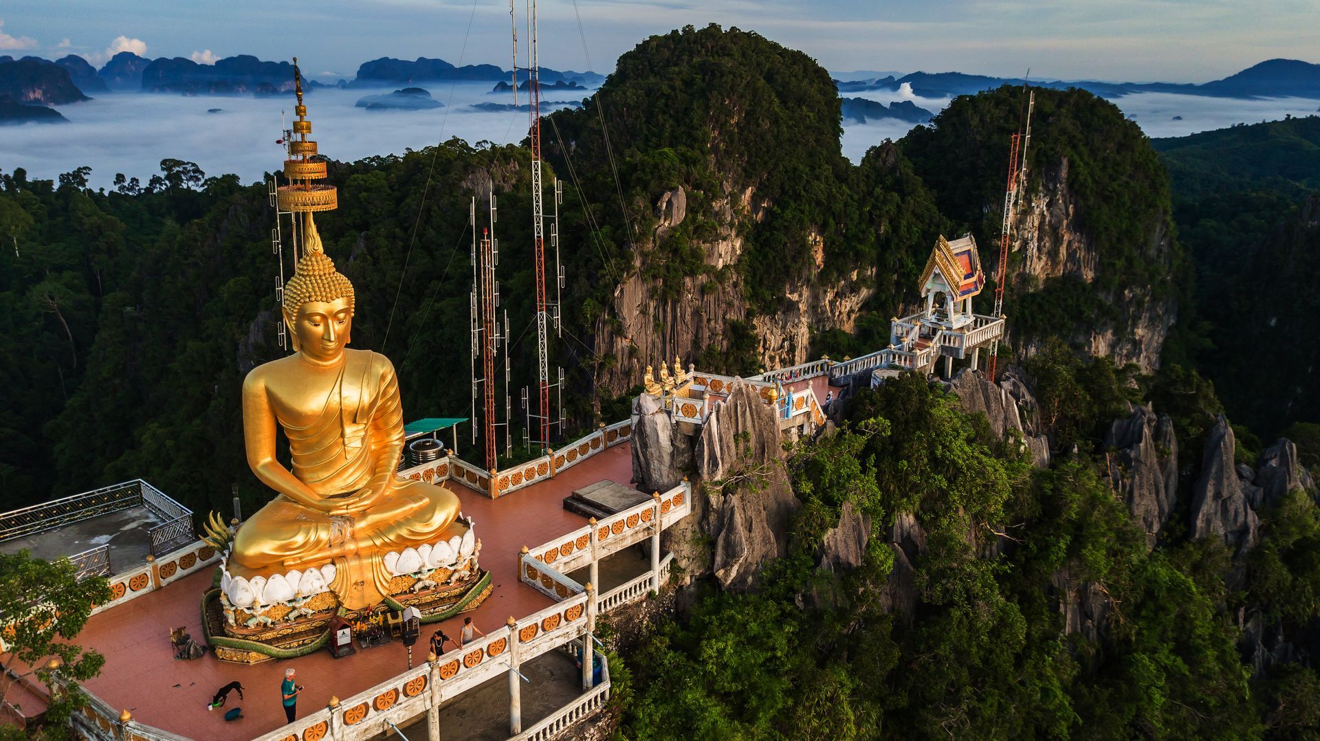 A large golden Buddha statue sits atop a rocky mountain peak overlooking a lush, misty valley in Thailand.