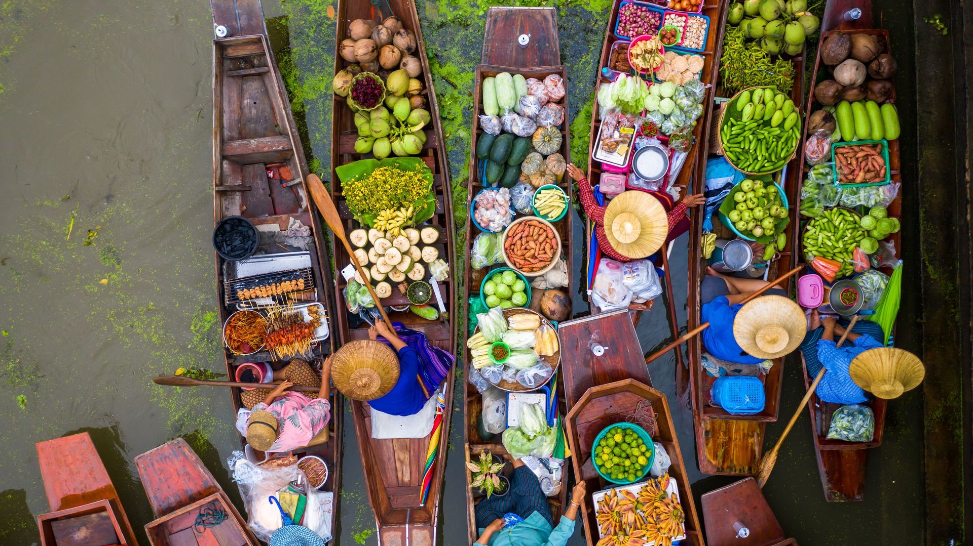 An aerial view of several wooden boats packed with colorful produce, flowers, and goods, floating on a canal market.