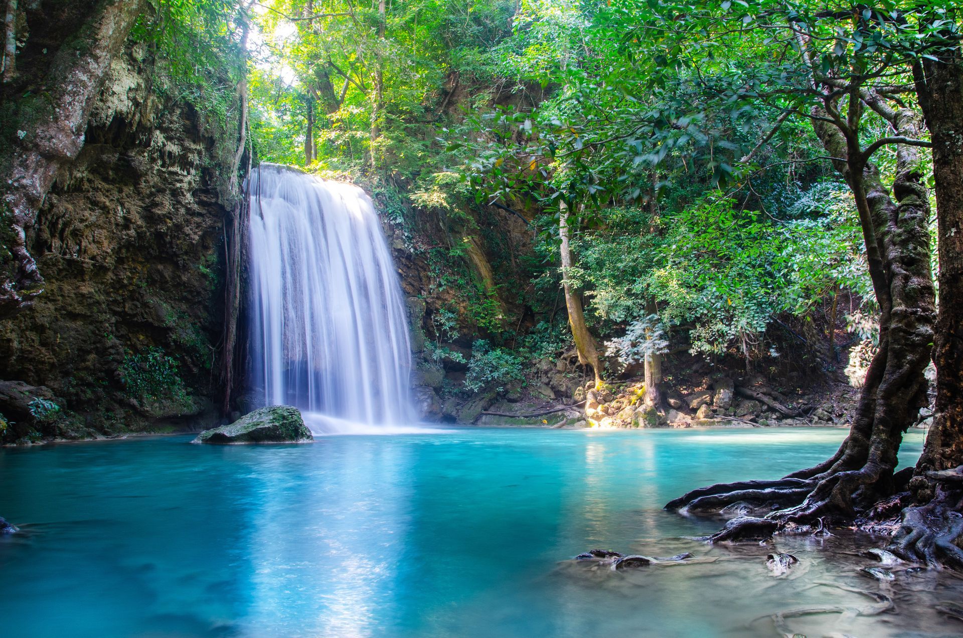 A vibrant, turquoise waterfall flows into a clear pool surrounded by lush green tropical trees and rocks.