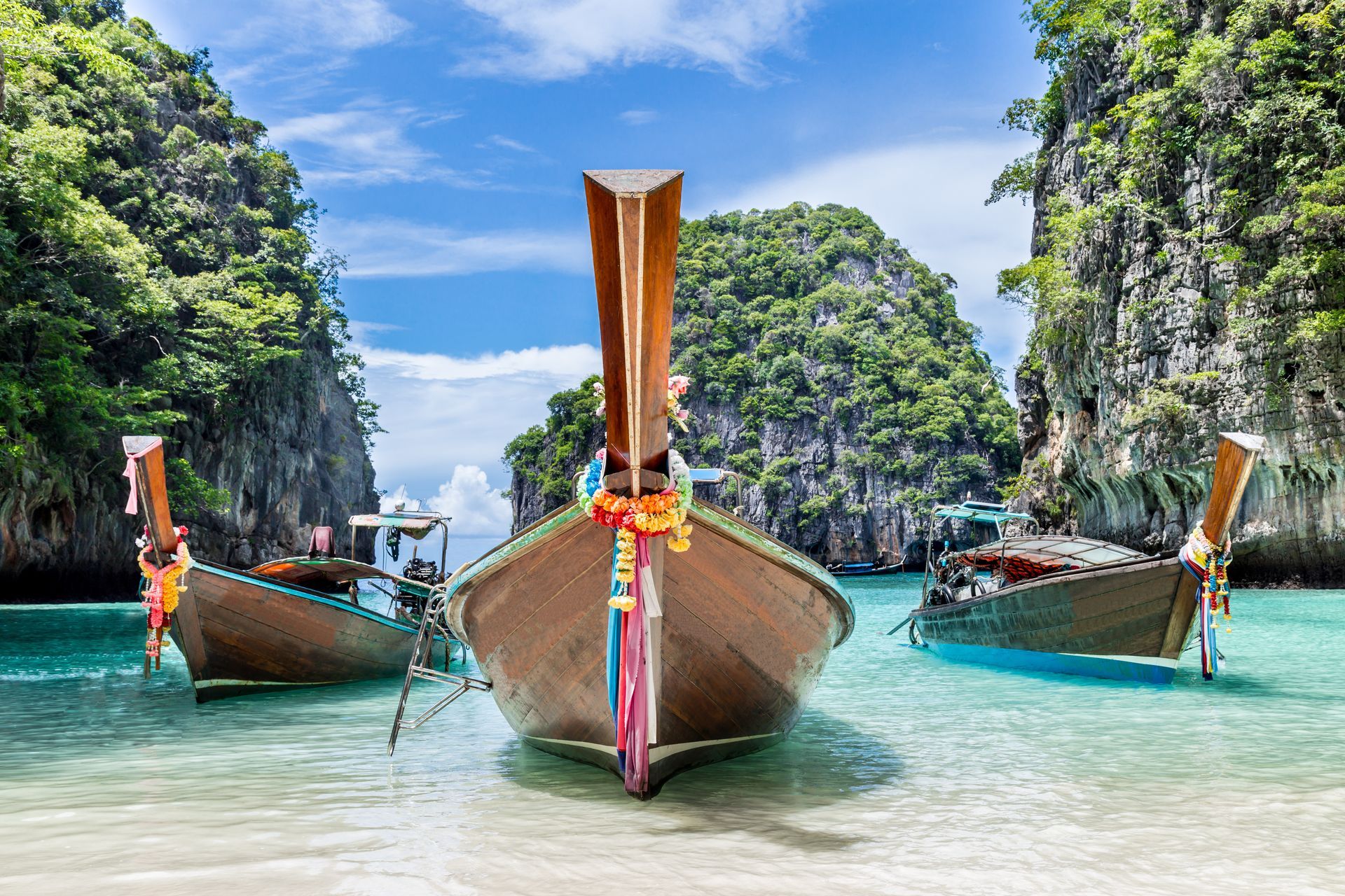 Three traditional wooden long-tail boats with colorful ribbons float in clear turquoise water between limestone cliffs.