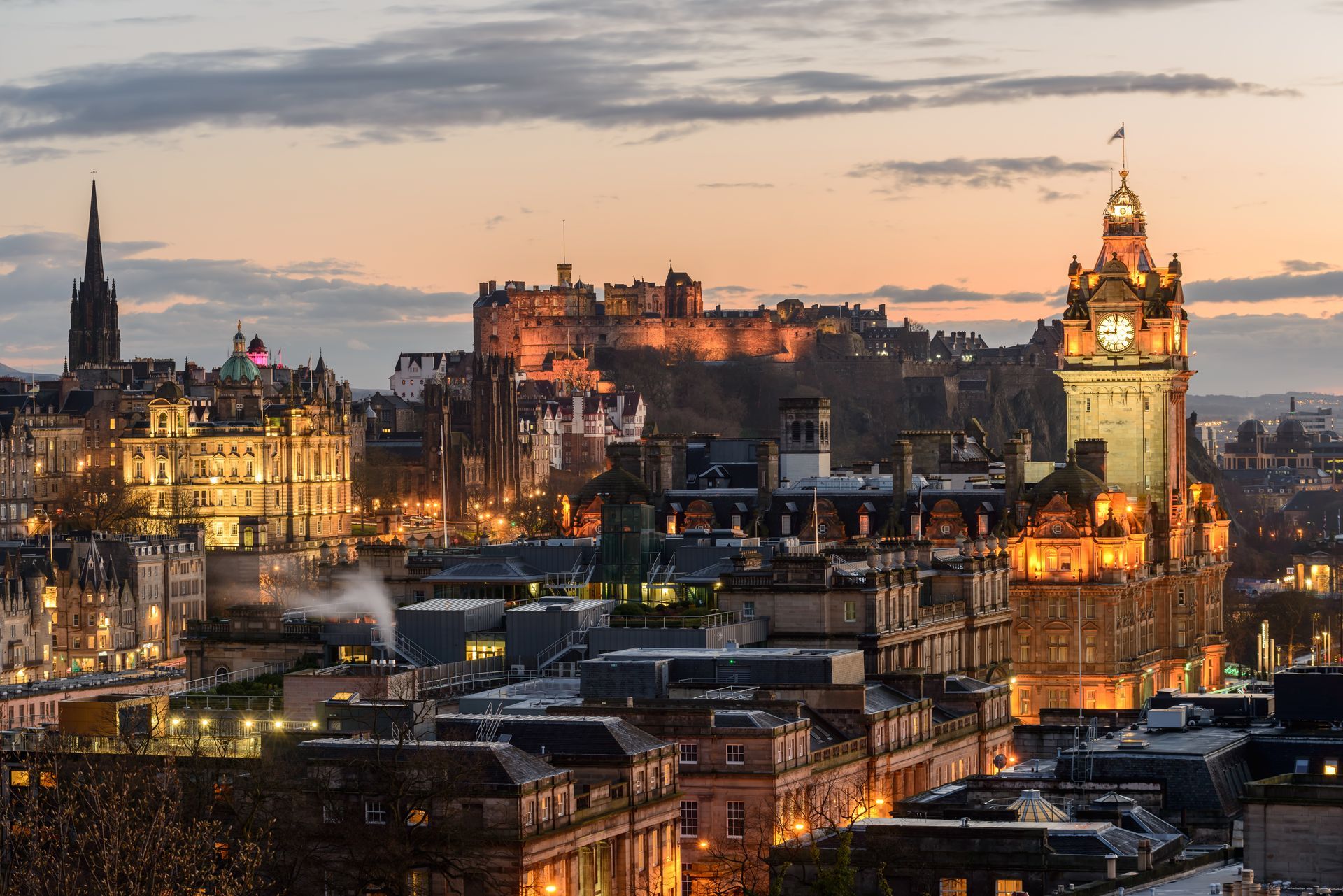 A golden hour view of Edinburgh Castle overlooking the city, featuring the illuminated Balmoral Hotel clock tower.