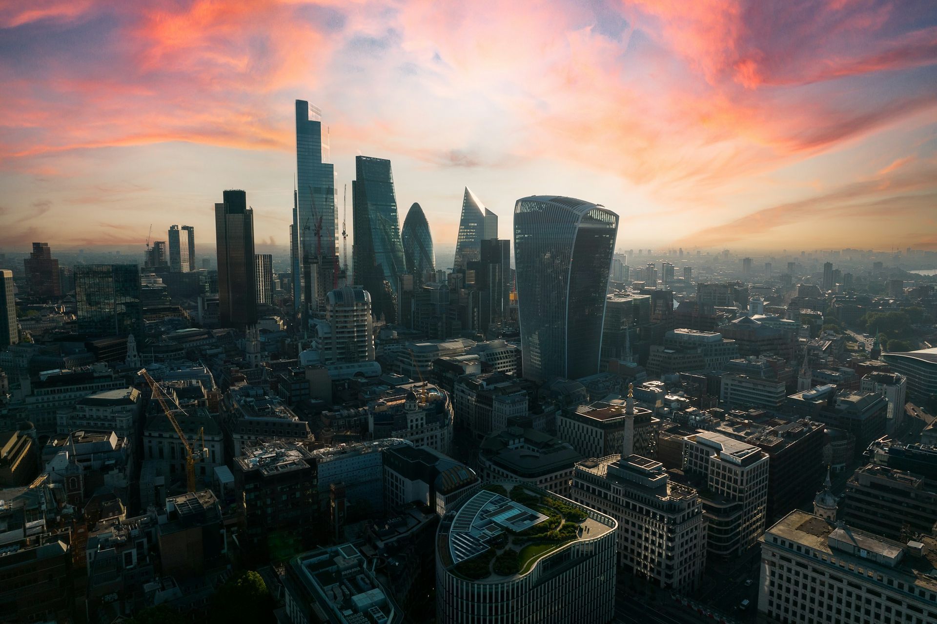 Aerial view of the London skyline featuring modern skyscrapers silhouetted against a vibrant sunset with pink clouds.