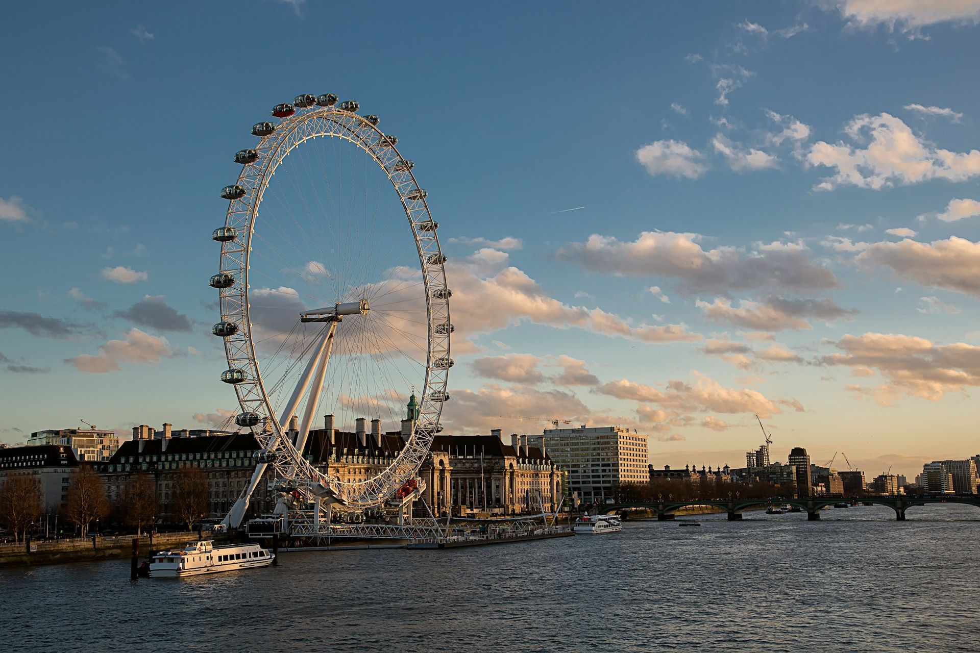 The London Eye ferris wheel stands on the banks of the River Thames in London under a soft, golden sunset sky.