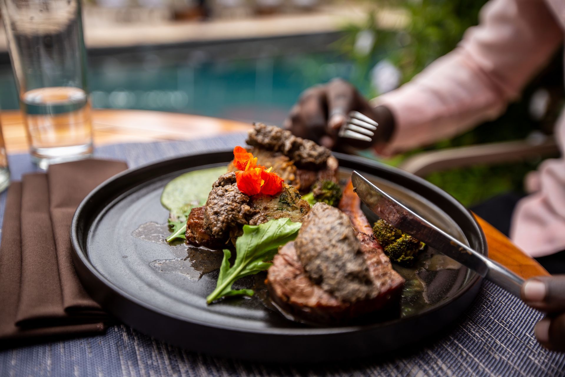 A person cuts a steak topped with herbs and greens on a black plate, with a glass of water nearby in an outdoor setting.