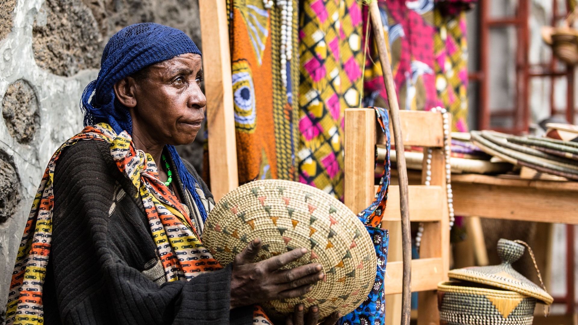 A person in a blue headscarf holds a woven basket among colorful textiles and wooden stools at an outdoor market.