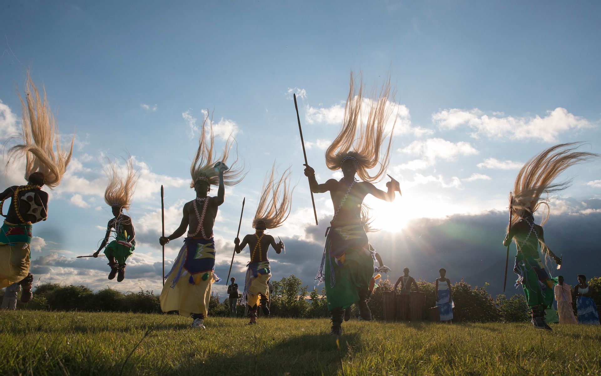 People in traditional attire jumping in a grassy field against a sunset sky with long, flowing headpieces.