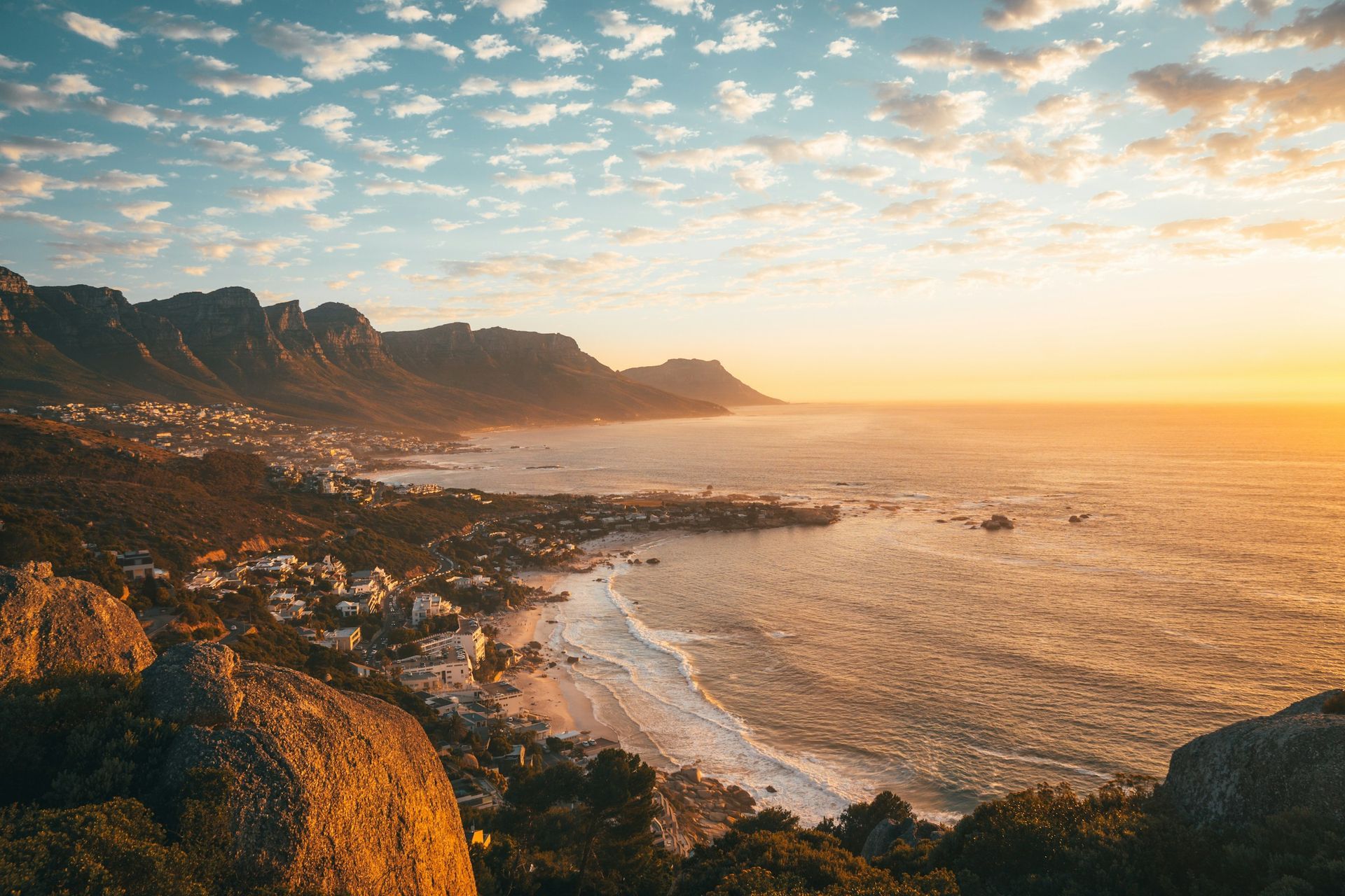 A scenic, sun-drenched coastal landscape featuring rugged mountains meeting the ocean at sunset with scattered clouds.