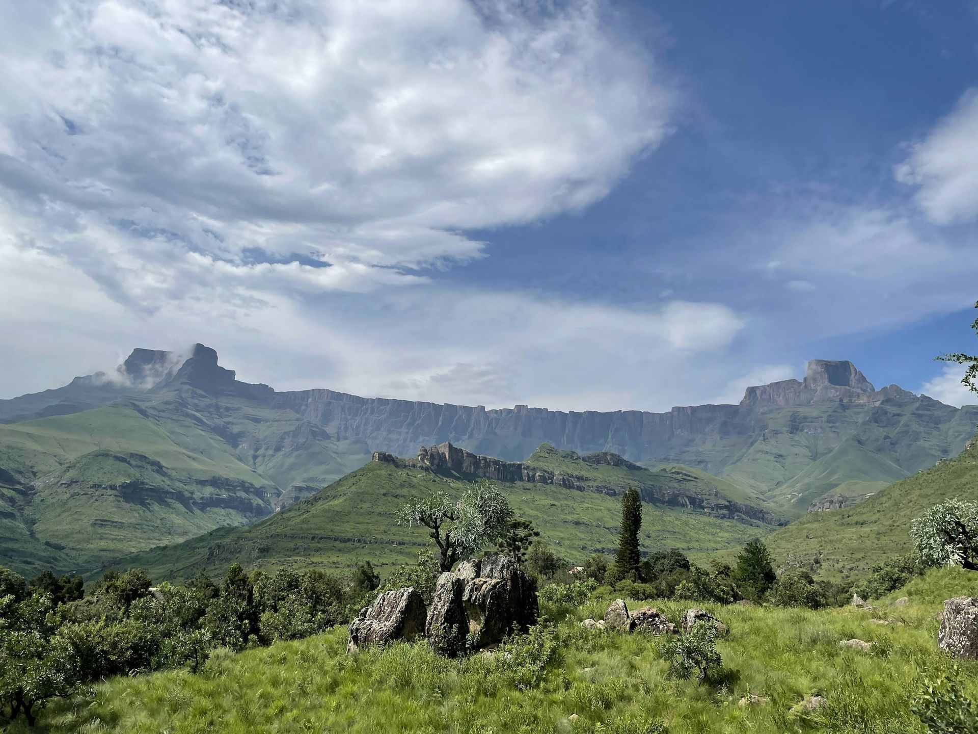 Lush green hills and jagged mountains under a blue, cloudy sky in the Drakensberg, South Africa.