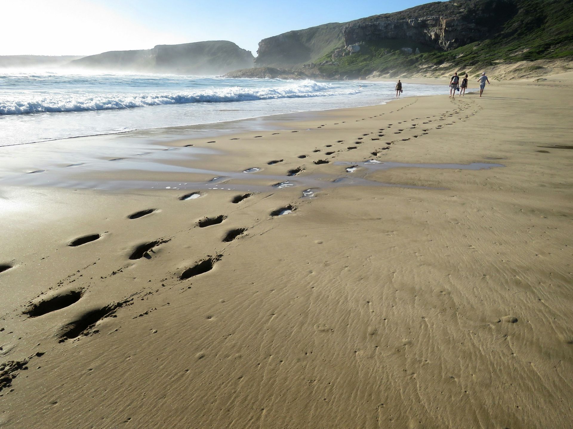 Footprints in the damp sand lead along a beach toward a group of people walking near the ocean waves and cliffs.