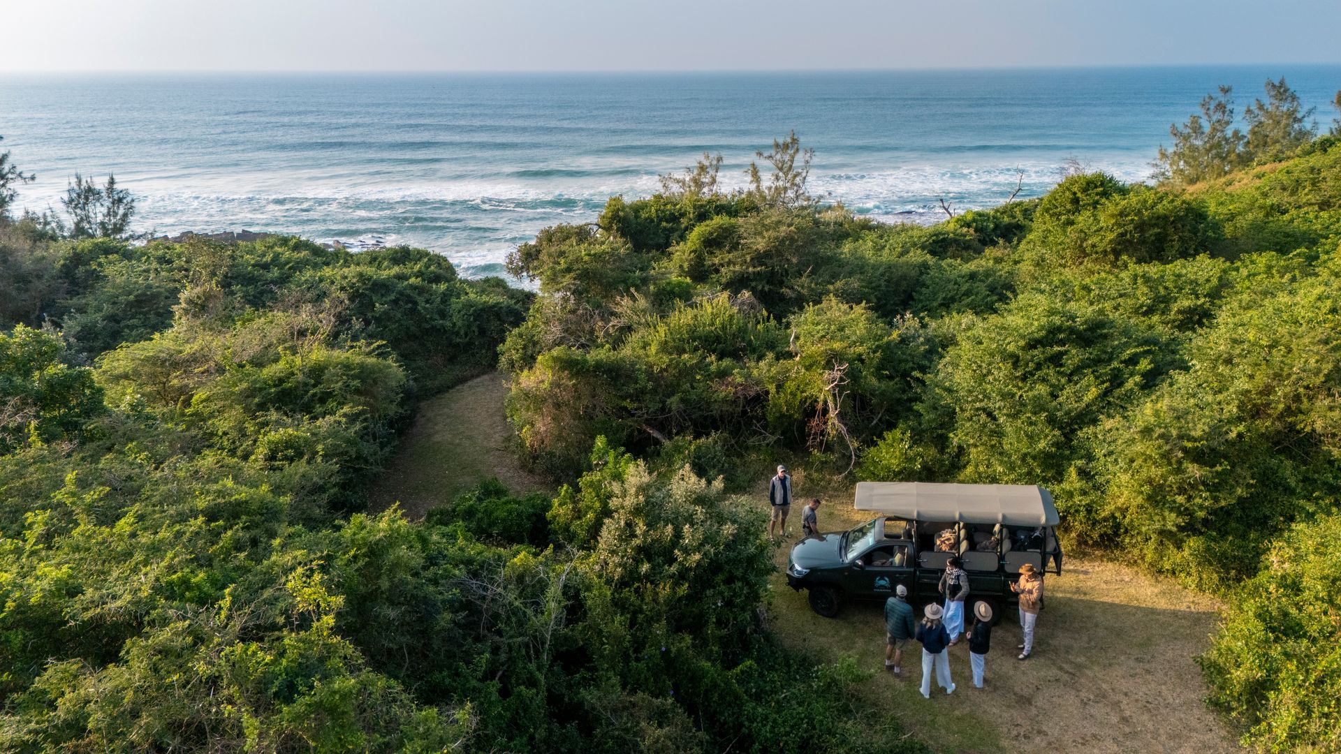 A group of people gathers near an open-air safari vehicle parked in lush coastal greenery overlooking the ocean.