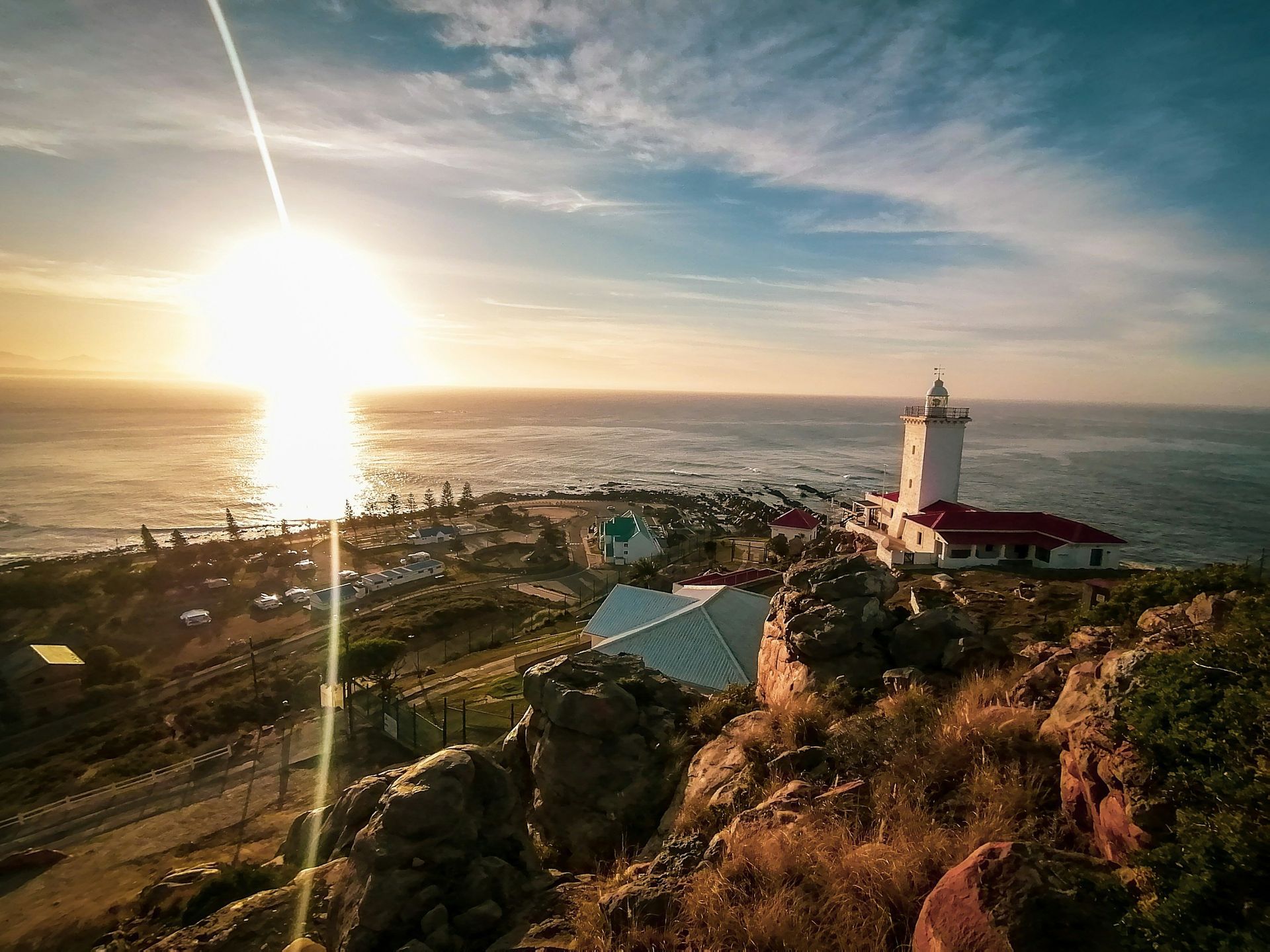 Lighthouse overlooking the ocean at sunset from a rocky, coastal hillside.