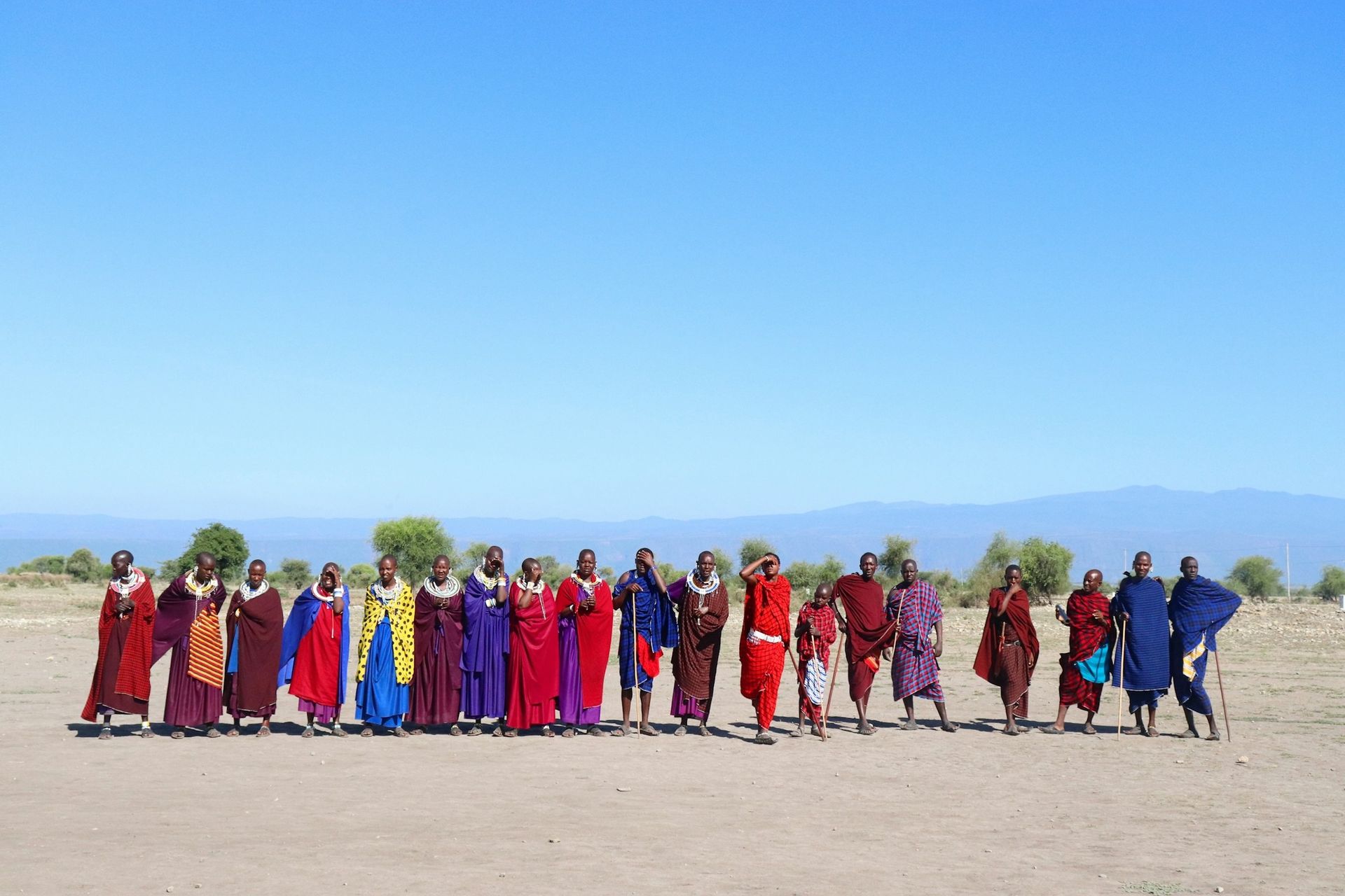 A group of people in colorful traditional shukas stand in a line on a dry, open plain under a bright blue sky.