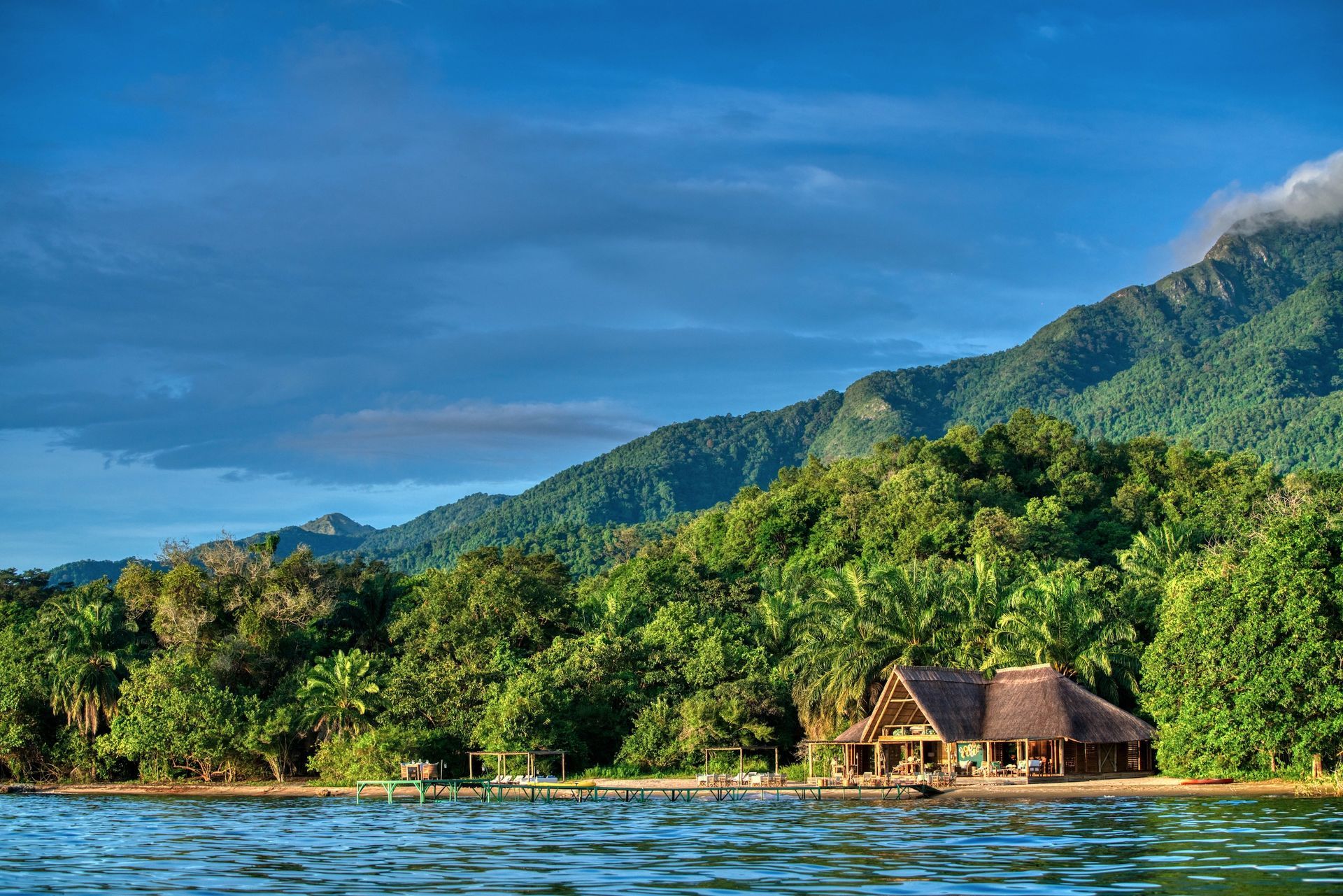 A wooden cabin with a thatched roof sits on a forested shoreline beneath lush, green tropical mountains under a blue sky.