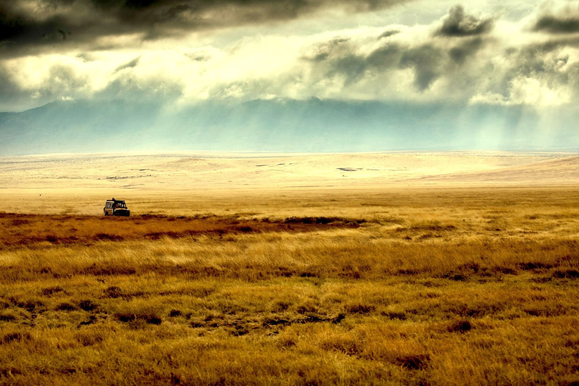 A lone vehicle travels through a vast, golden savanna under a dramatic, cloudy sky with sunbeams breaking through.
