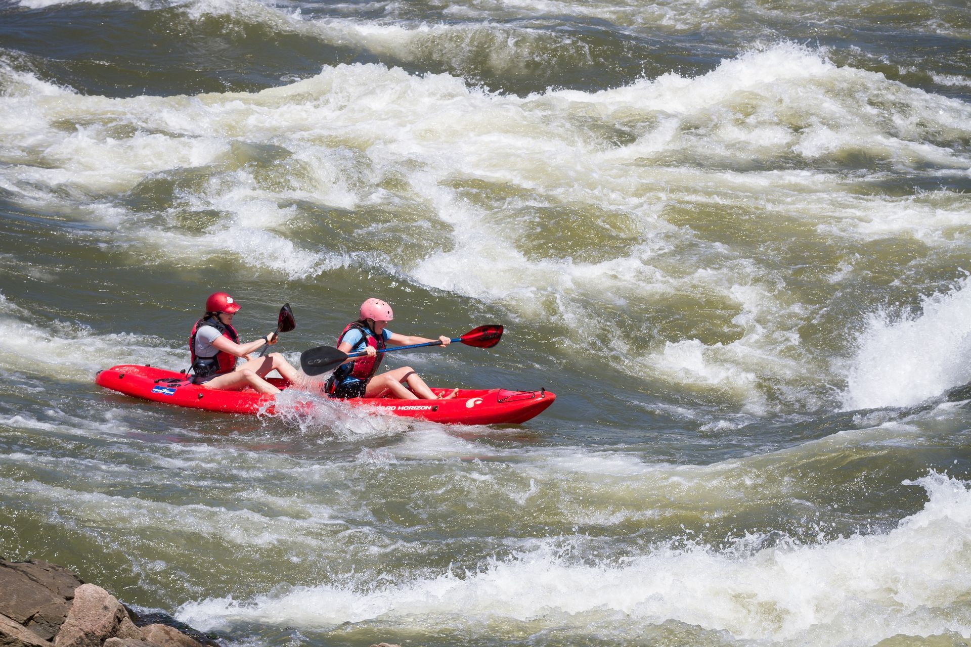Two people in life vests and helmets navigate a red tandem kayak through turbulent river rapids.