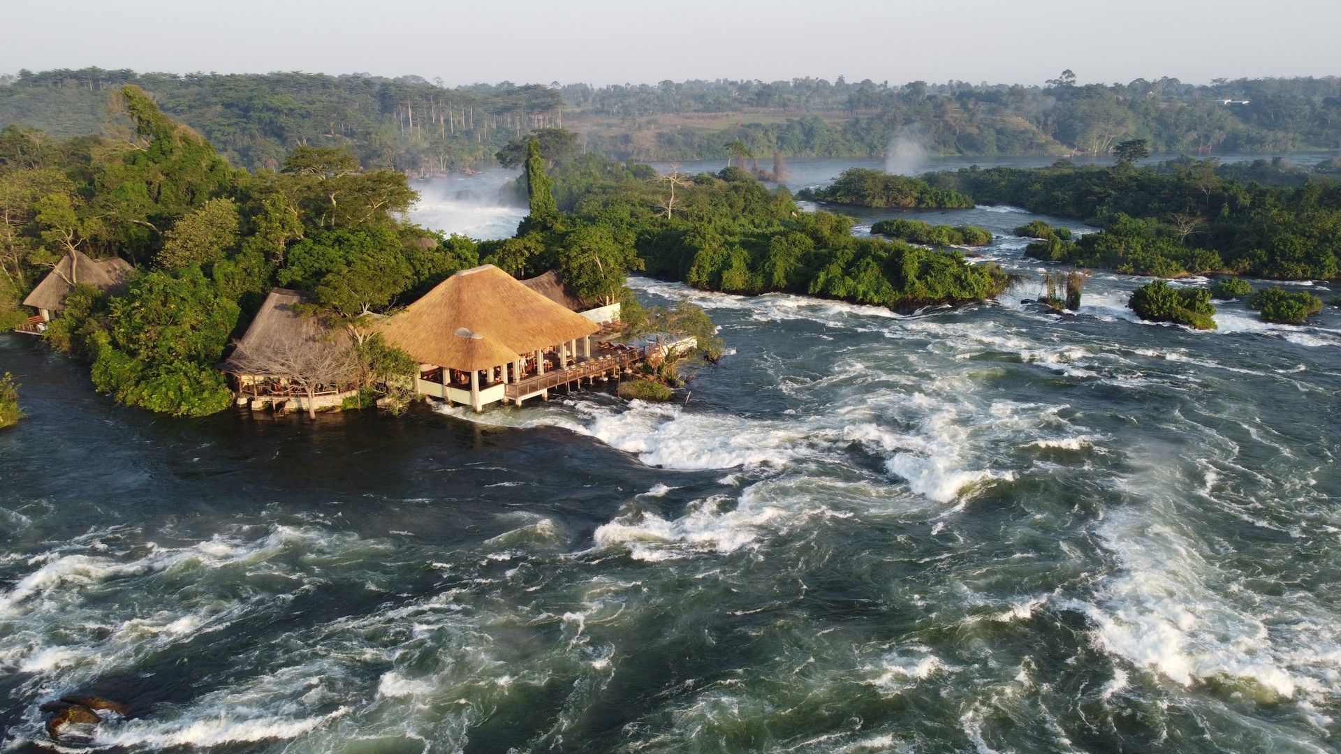 A thatched-roof building sits on a lush island surrounded by the rushing white water of the Nile River at Bujagali Falls.