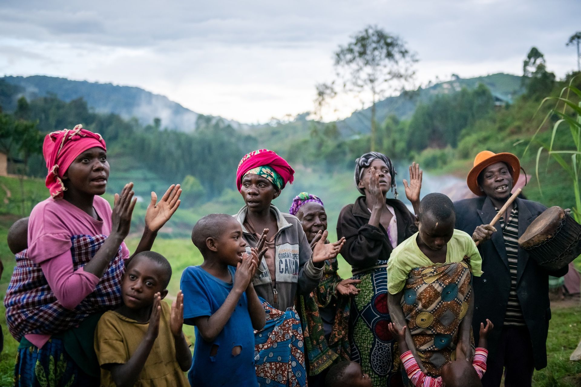A group of people singing and clapping outdoors in a hilly, rural landscape, with one person playing a small drum.