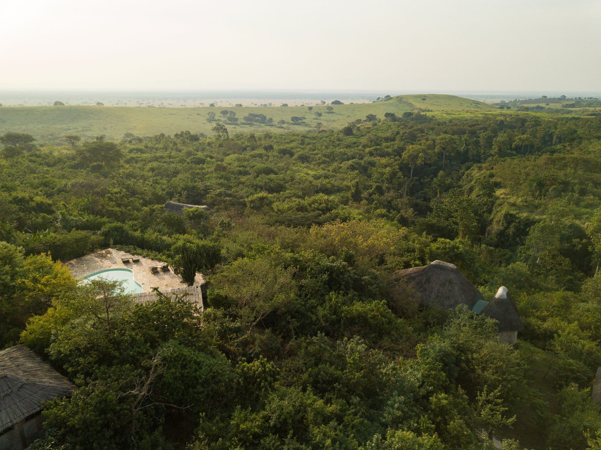 Aerial view of a secluded lodge with a swimming pool nestled in lush green forest overlooking a vast savanna landscape.