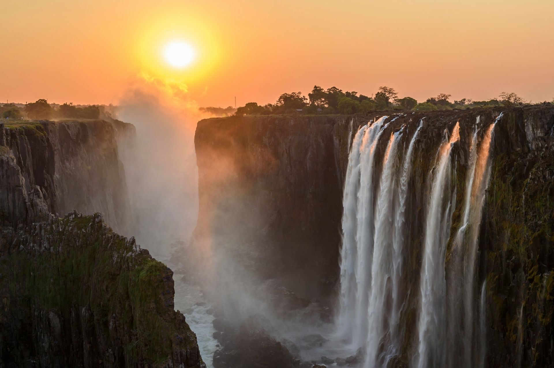 Victoria Falls at sunset, with golden light illuminating cascading water and mist rising from the gorge.